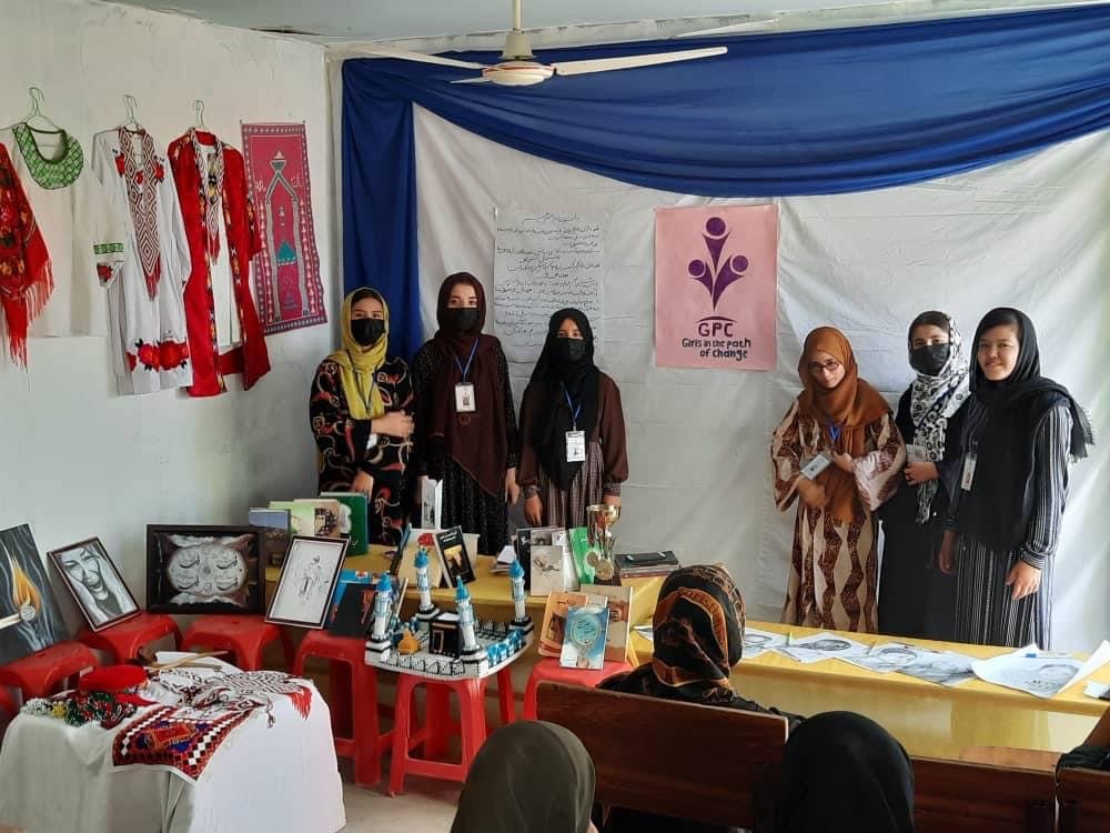 A group of six women standing in a room with cultural decorations, display tables with artwork and crafts, and a banner that reads 'Girls in the pocket of change.' They are wearing masks and traditional and modern clothing.