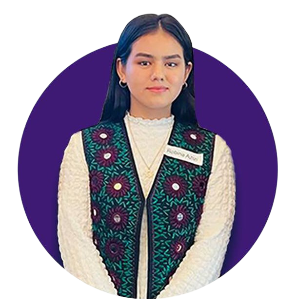 A woman with dark hair wearing a cream-colored sweater and a colorful vest with floral patterns, standing in front of a purple background.