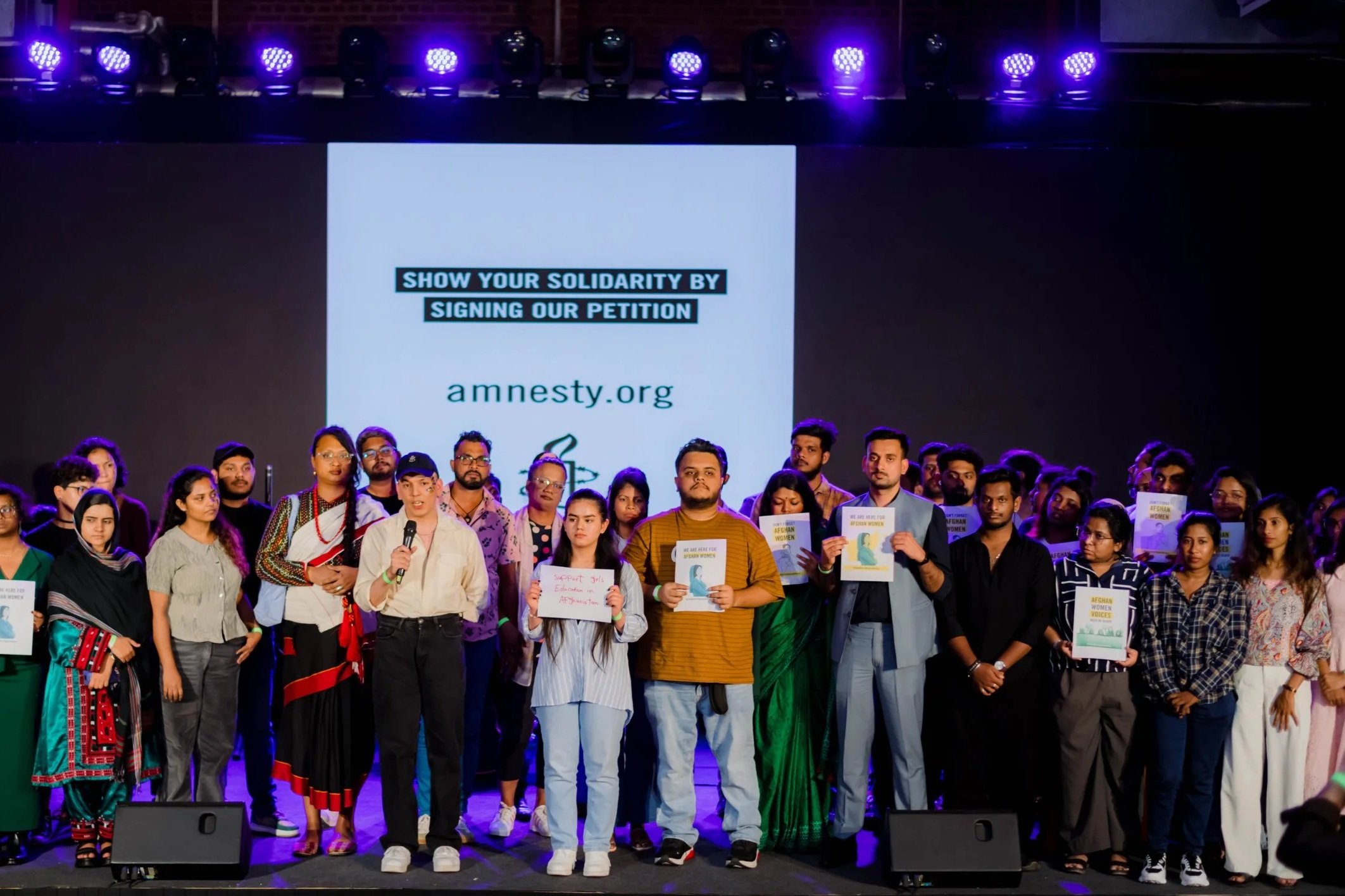 A diverse group of people gathered on stage at an event promoting solidarity with Afghanistan, holding signs and posters, with a large screen in the background displaying a message to show support by signing a petition on amnesty.org.
