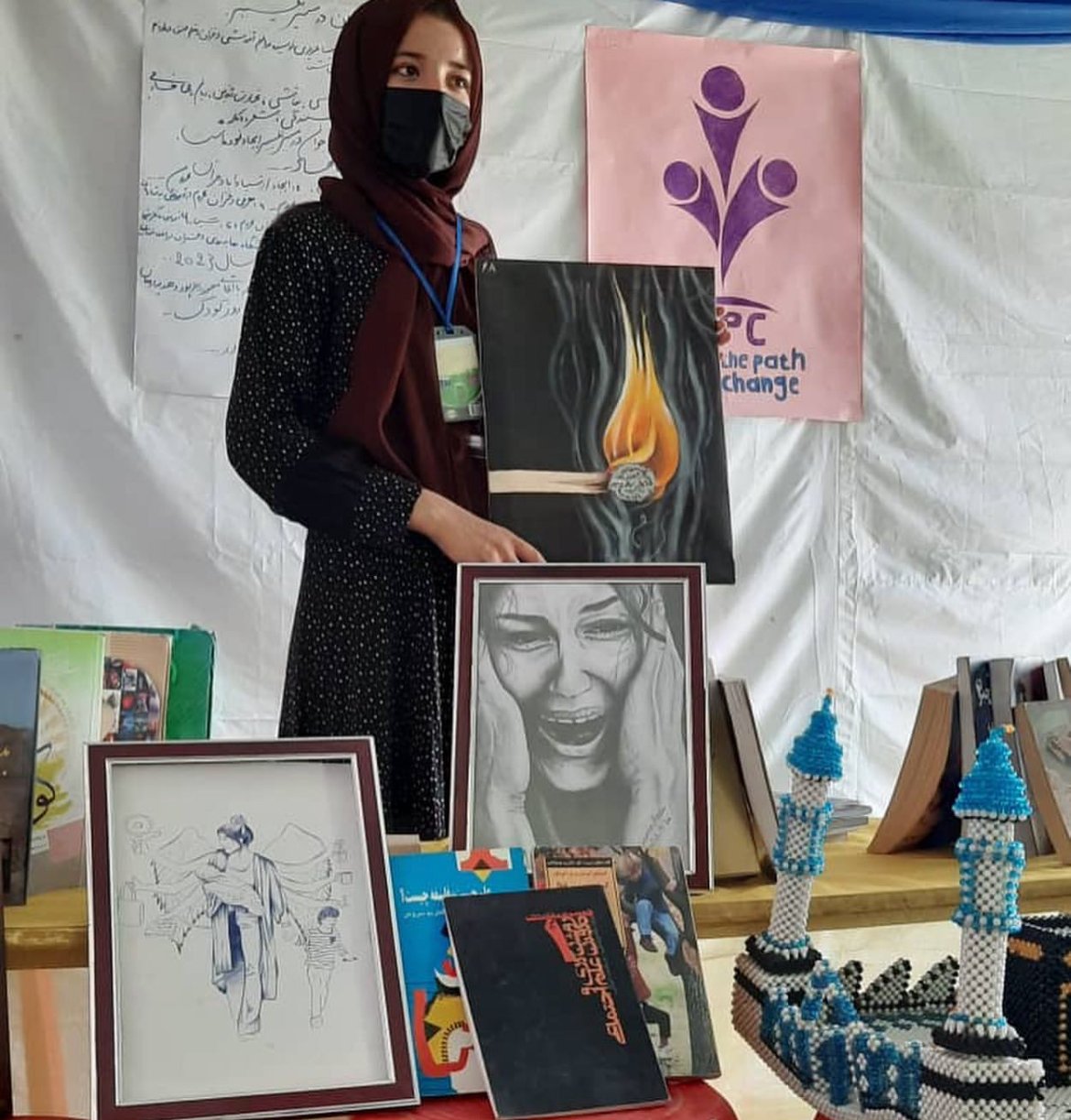 A woman in a black mask and maroon headscarf stands behind a display table of artwork and crafts at an indoor event. She is holding a painting of a matchstick with a burning flame and a pencil drawing of a crying woman. The background has a pink banner with purple logo and text, and various framed artworks and bead crafts are on the table.