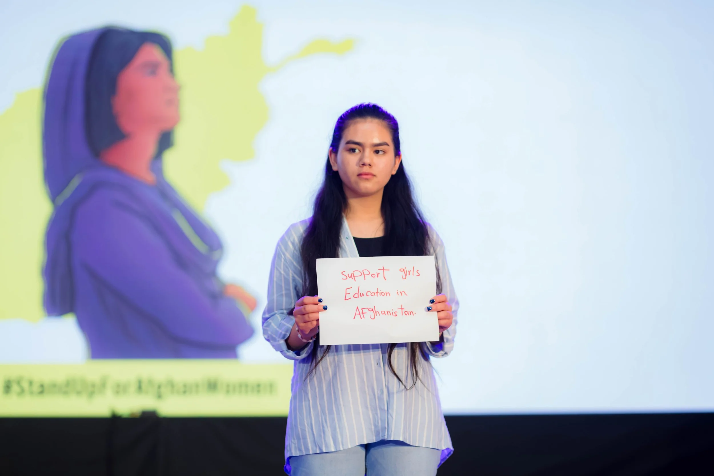 A young woman with long dark hair wearing a striped shirt holding a sign that reads 'Support girls' Education in Afghanistan' during a presentation or protest. A blurred figure of another woman is visible in the foreground.