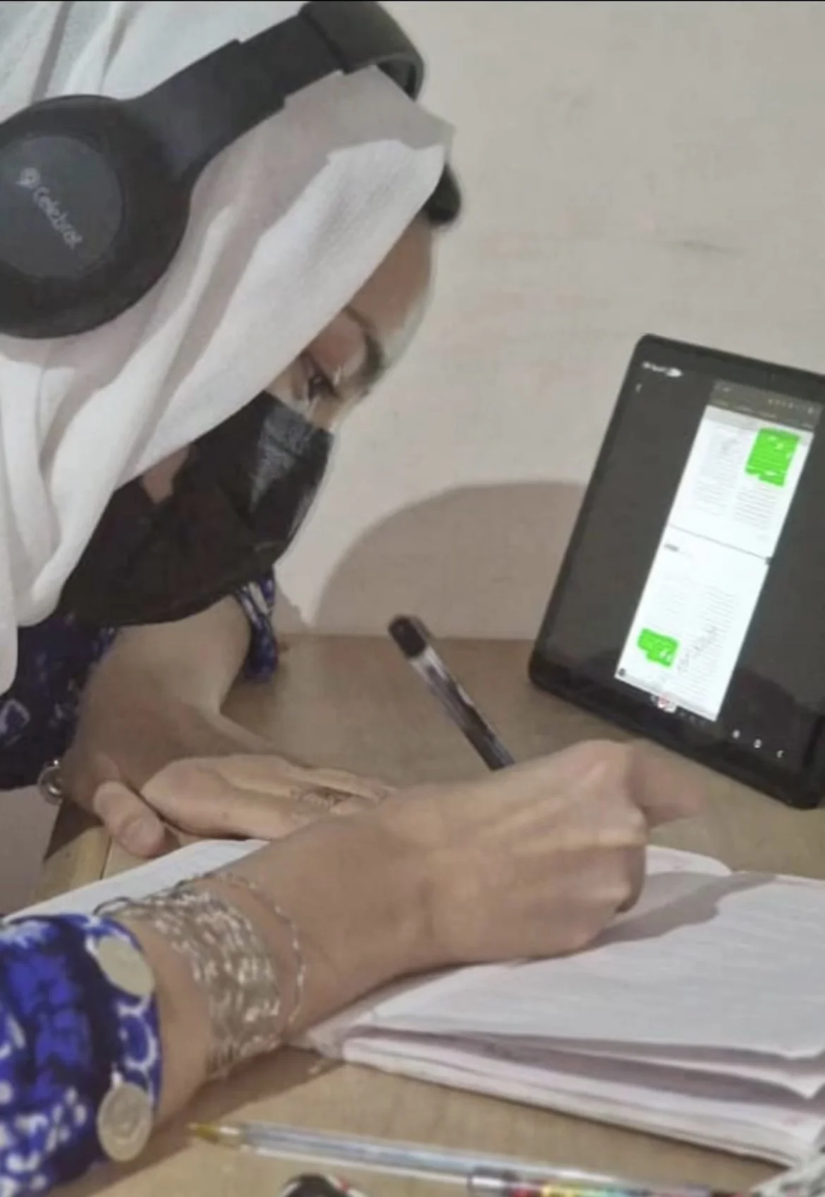 A person wearing a protective face mask, black shirt, and purple patterned sleeves is sitting at a desk, writing in a notebook with a black pen. There is a tablet computer on the desk displaying a document with green highlights and a pen nearby.