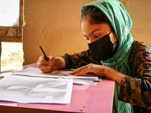 A woman wearing a green headscarf and black face mask sitting at a pink table, writing on papers with a pen.