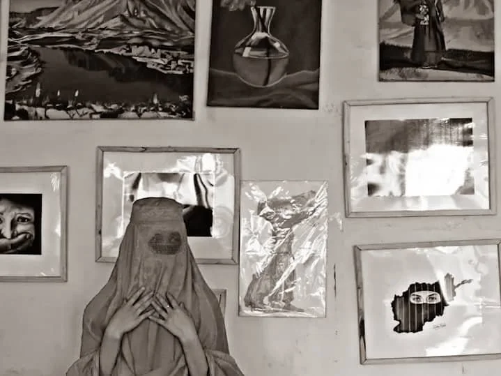 A person is wearing a cloth over their head, resembling a bridal veil, and is holding their face with their hands, standing in front of a wall decorated with various framed artworks.