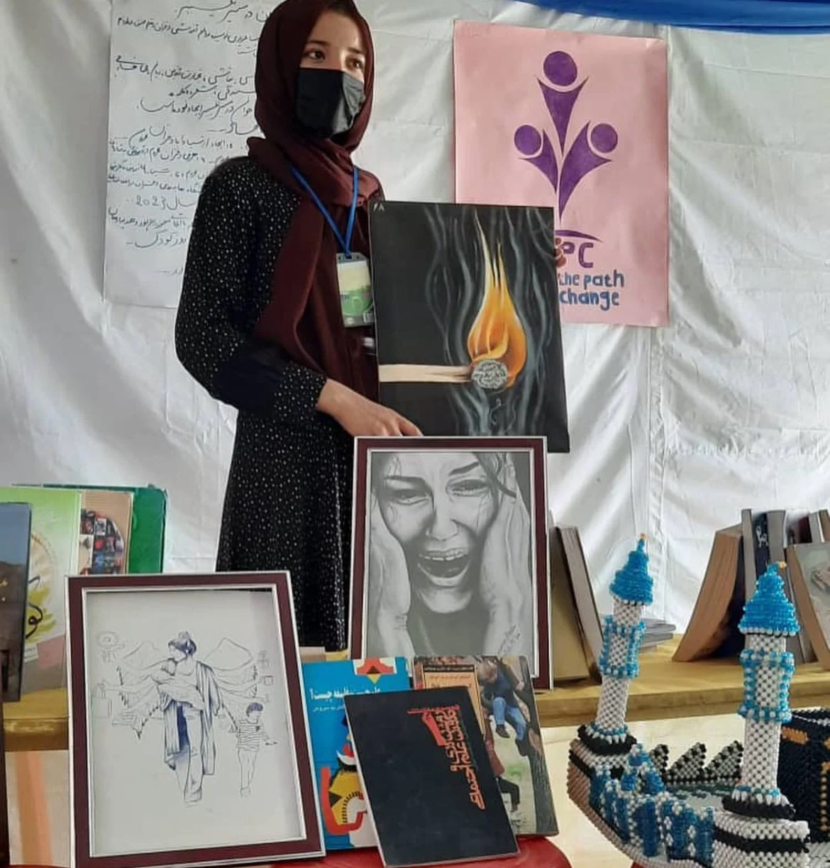 A woman wearing a black face mask and a dark red hijab holds a black canvas painting of a matchstick with a flame, burning a gray ball of paper. She stands in front of a white fabric backdrop with posters and artwork on display, including a black-and-white sketch of a joyful woman, beaded models resembling towers, and various books and art pieces on a table.