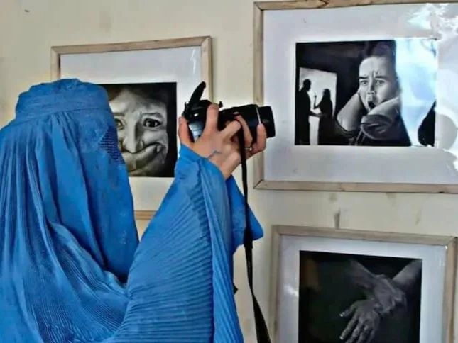 Person dressed in blue holding a camera and taking a photo of black and white framed photographs on a wall.
