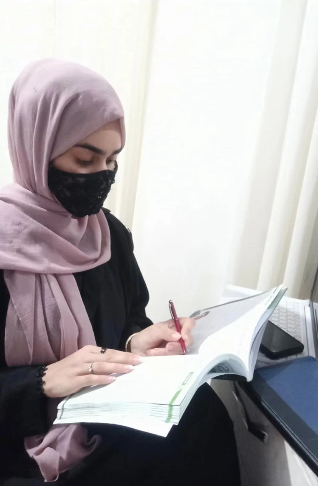 A woman with a pink hijab and a black face mask is reading a book and taking notes with a red pen at a desk.