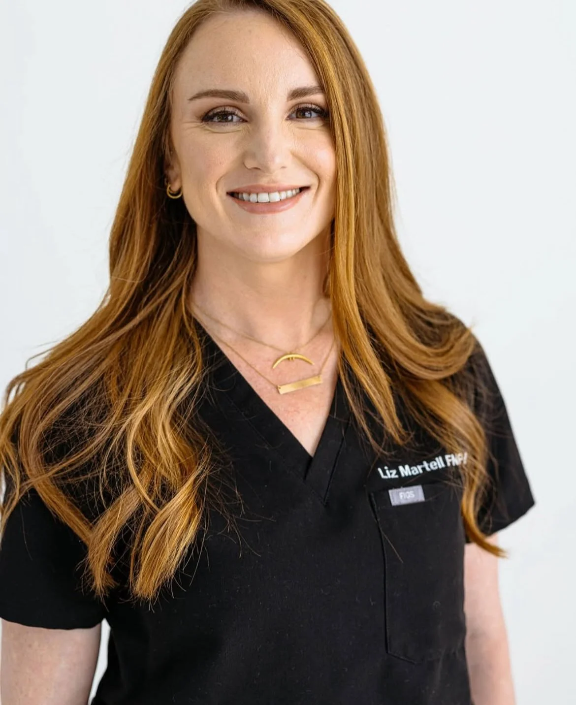 A woman with long red hair, wearing a black uniform and gold jewelry, smiling at the camera.
