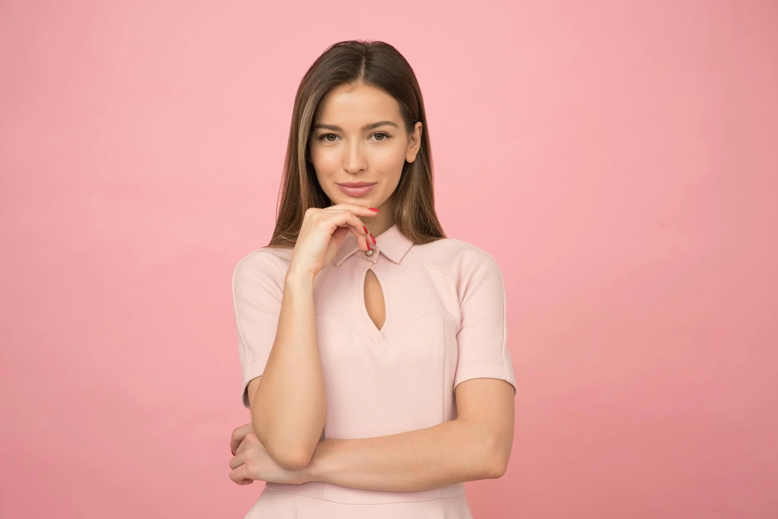 A young woman with long brown hair wearing a light pink top against a pink background. She is smiling softly, with one hand resting near her chin and the other arm crossed.