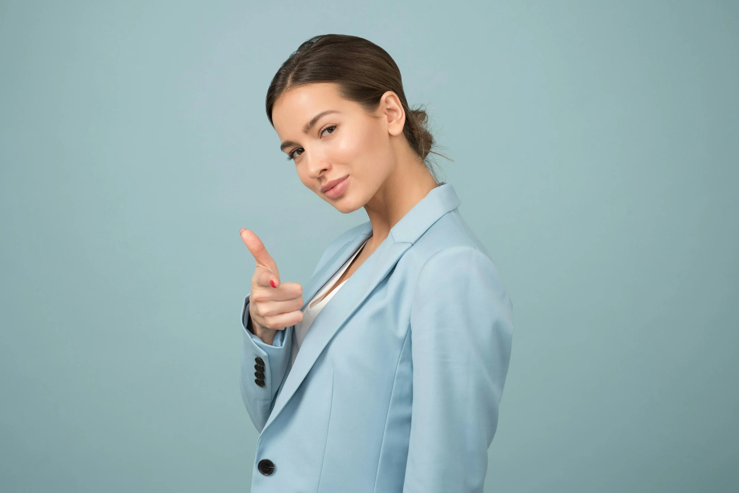 A young woman with brown hair in a low ponytail, wearing a light blue blazer, points with her index finger while smiling softly against a plain teal background.