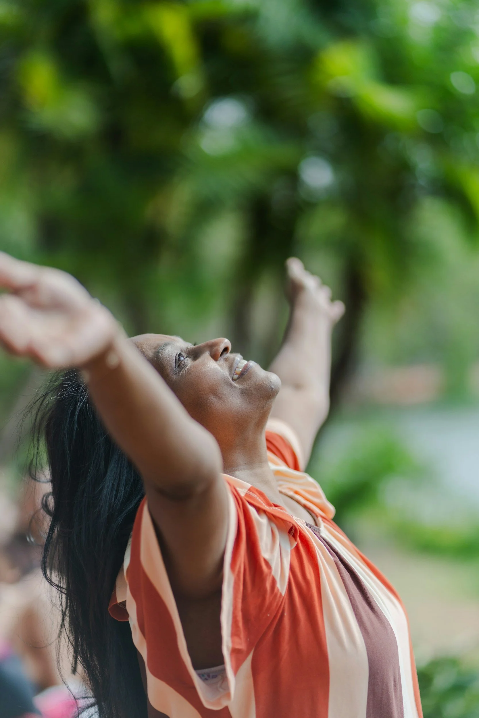 A person smiling outdoors with arms raised, surrounded by green trees.
