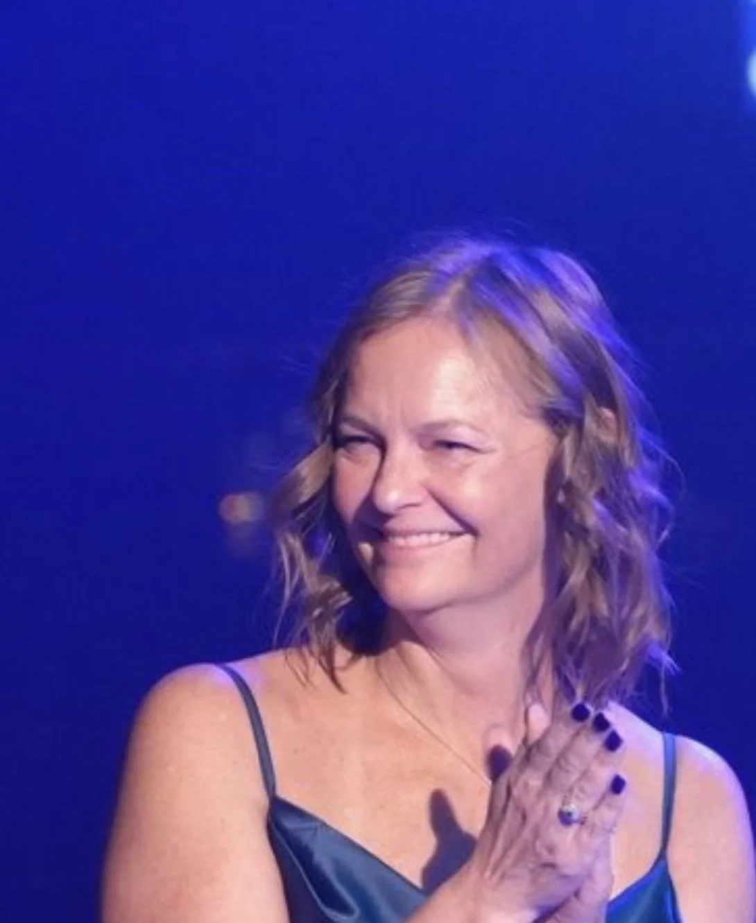 A woman with wavy blonde hair wearing a black spaghetti strap dress, smiling and clapping in front of a blue background.