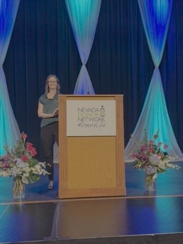 A woman standing behind a wooden podium with the Nevada Network logo, on a stage decorated with flowers and blue draped curtains with colorful lighting.