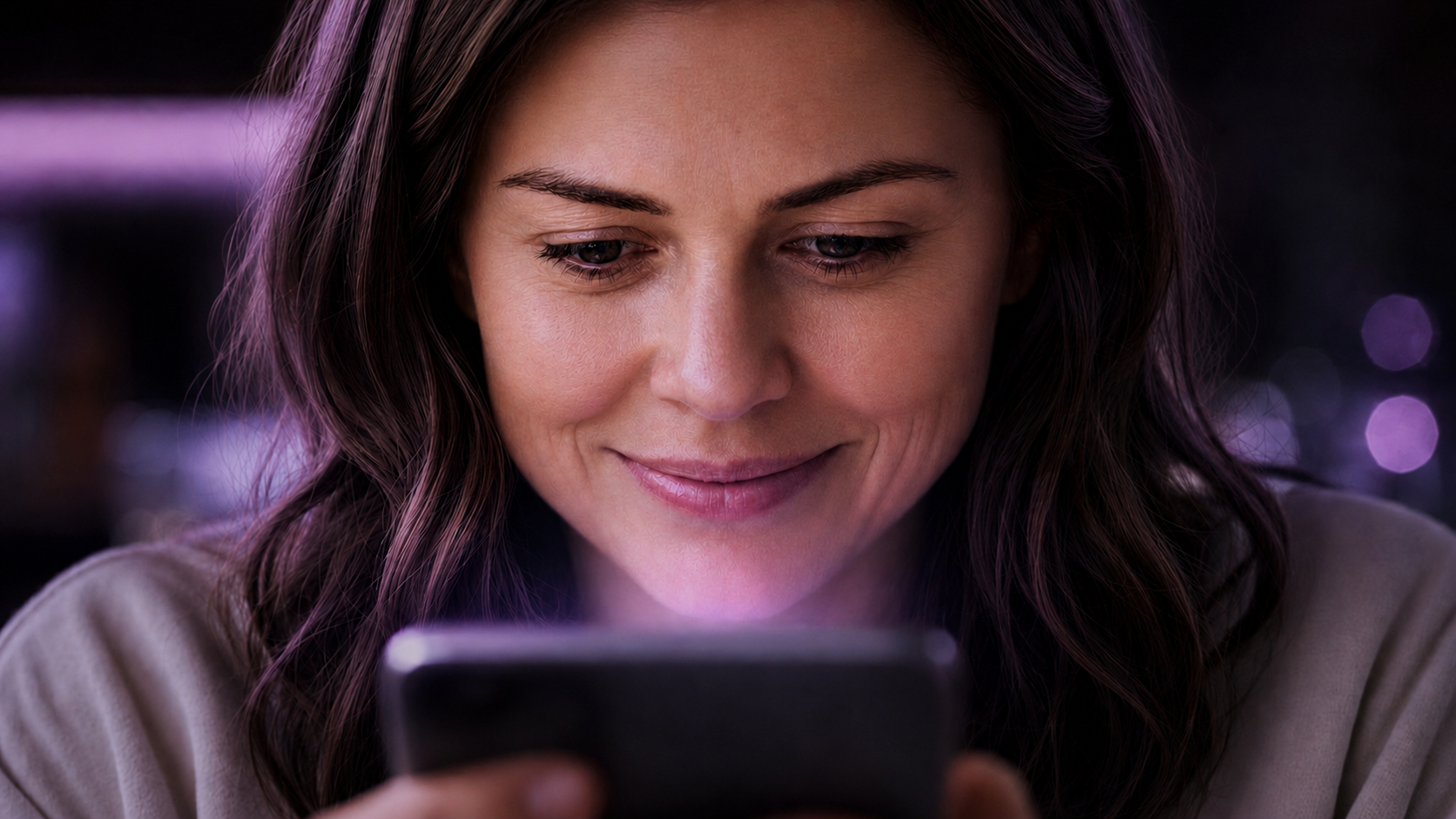 A woman with long brown hair smiling while looking at her phone in a dimly lit environment with purple and pink bokeh lights in the background.