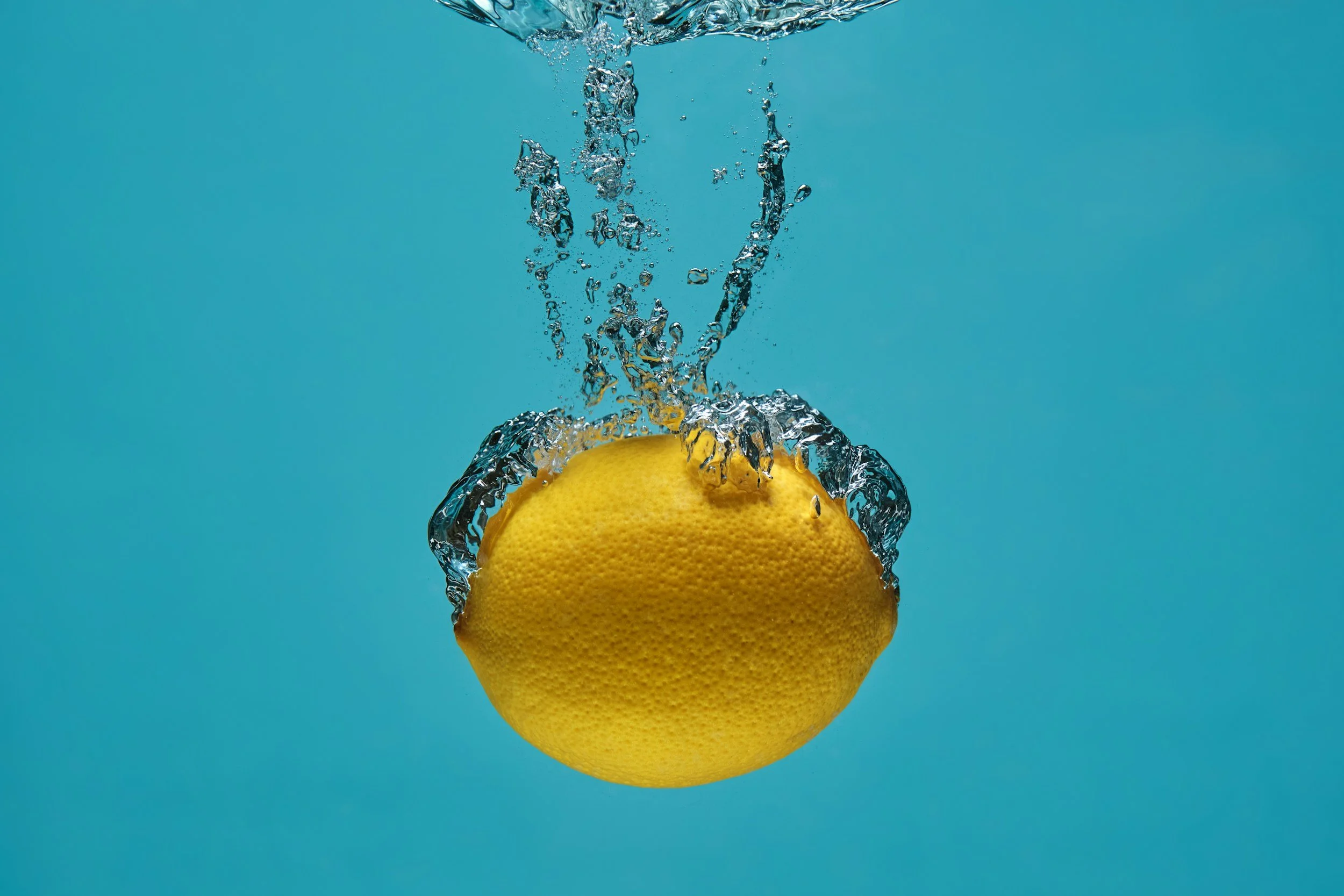 Lemon being dropped into water, creating splash and bubbles against a blue background.
