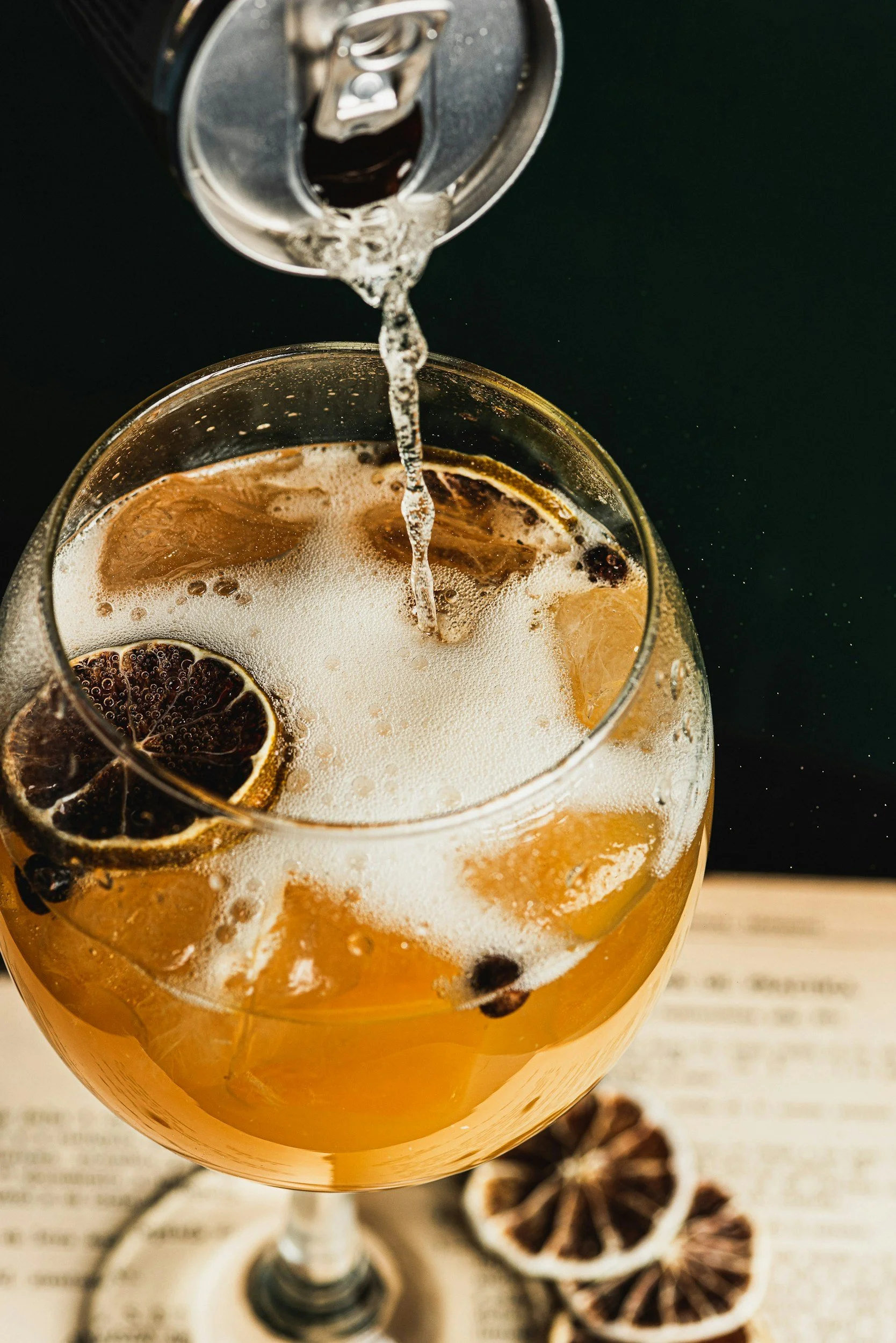 Close-up of a glass of iced drink with lemon and black tea, being poured from a soda can, with dried lemon slices and star anise around the glass on a paper background.