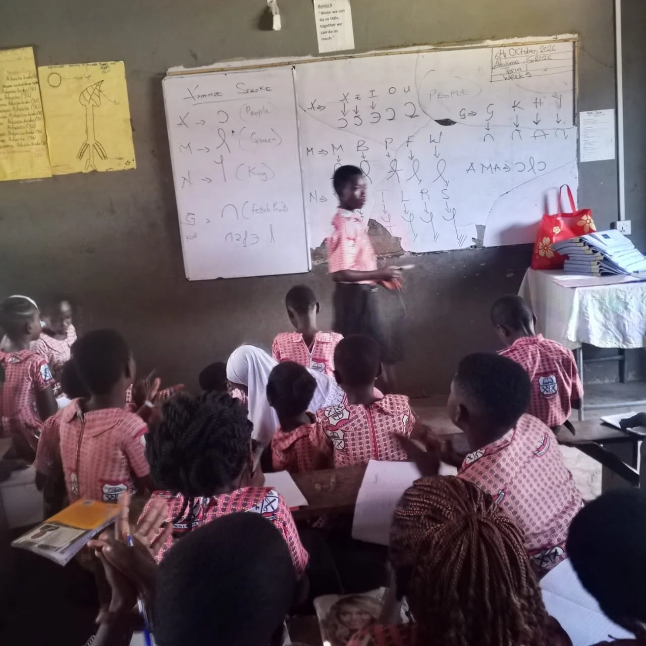 A classroom with young students wearing red and white checkered uniforms, seated at desks, listening to a teacher at the whiteboard. The teacher is standing in front of the class, explaining something written on the whiteboard. There are stacks of papers and a red bag on a table to the side, and various posters on the wall.