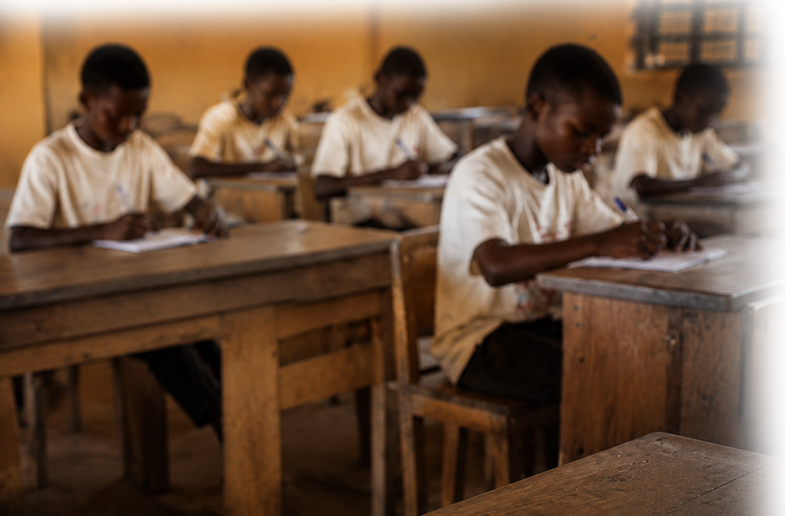 Young students seated at wooden desks in a classroom, writing in notebooks.