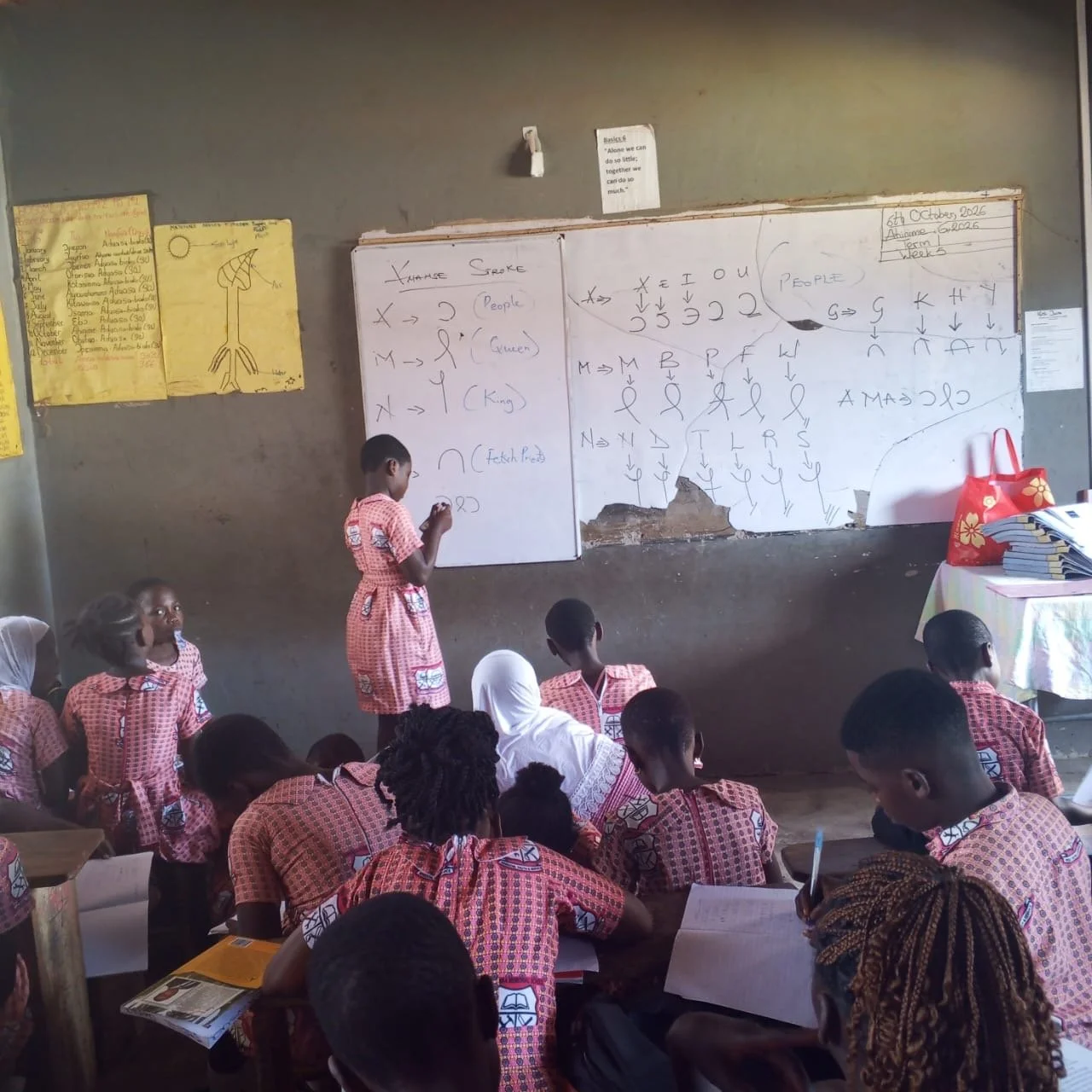 A classroom scene with students wearing red checkered uniforms, seated on the floor, with one student standing and writing on a whiteboard filled with chalk marks and symbols. There are posters on the wall and a table with a red bag and stacks of books.