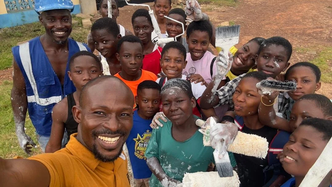 Ananse Stroke Foundation Teachers and students, smiling, holding paintbrushes with paint, outdoors after painting Ananse a new home.