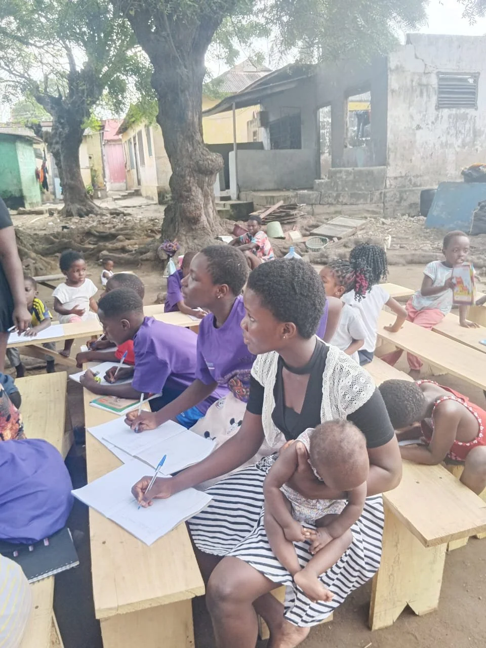 Children and women seated on wooden benches outdoors, some writing in notebooks, with large trees and houses in the background.