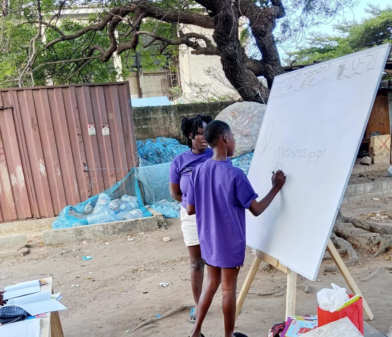 Two children wearing purple Ananse Stroke t-shirts are writing on a whiteboard outdoors in a dirt area with trees and collected plastic bottles in the background. One child is writing while the other watches. Ananse Stroke ghetto operations.