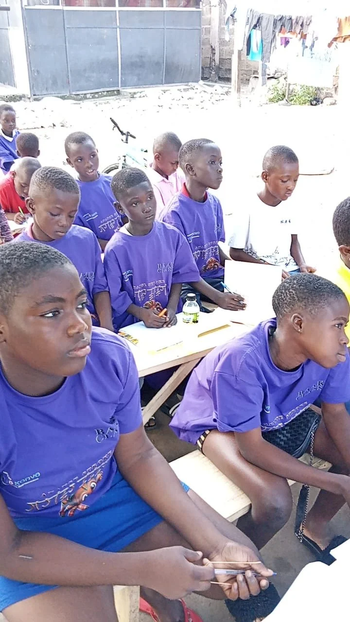 Group of young boys sitting at desks outdoors, some wearing purple Ananse Stroke t-shirts, in a classroom setting. Learning the Ananse Stroke