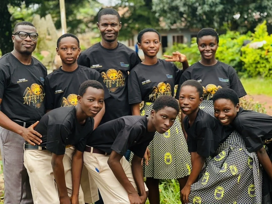Group of nine smiling children and teenagers outdoors, wearing matching black T-shirts with a bee graphic and another with a yellow and black patterned skirt, posing for a photo with a lush green background.