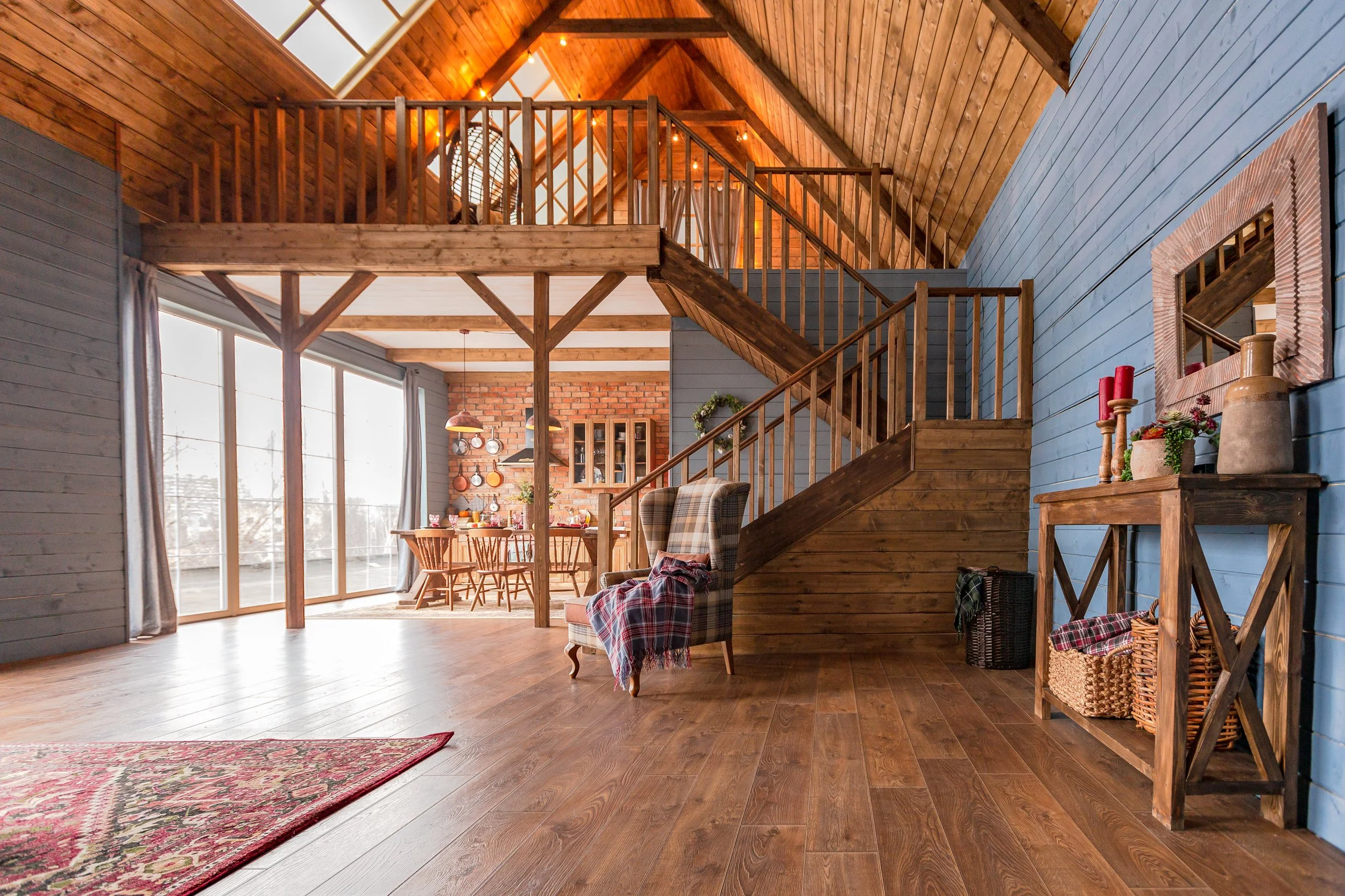 Interior view of a loft-style living room with wood-paneled walls, a staircase, a dining area, and cozy seating, illuminated by large windows and warm lighting.