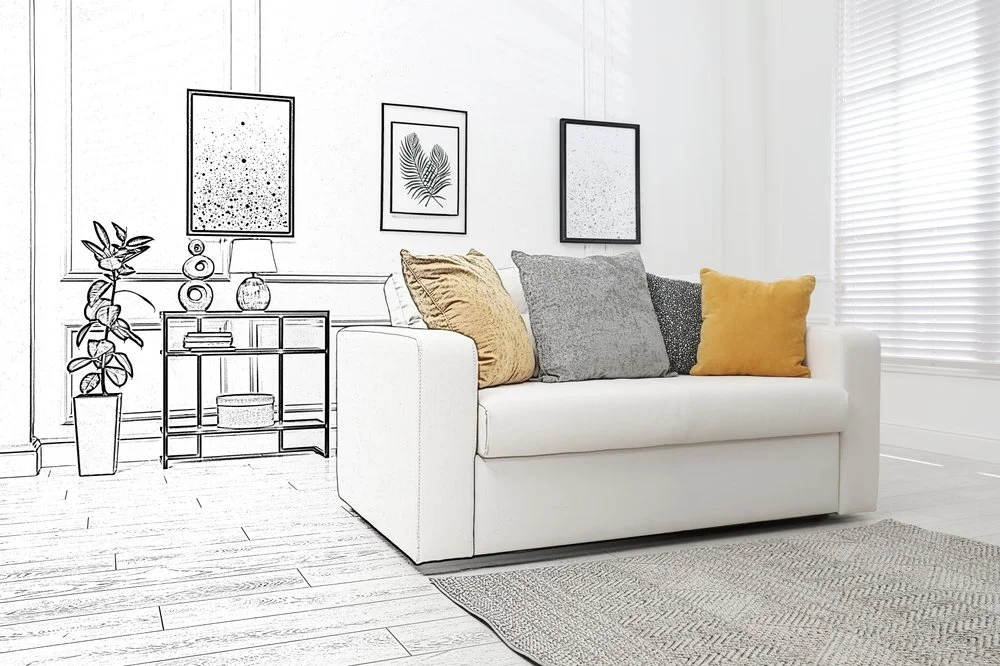 A living room with white walls, a white couch with yellow and gray pillows, a side table with a lamp, framed art on the wall, a potted plant, and window blinds.