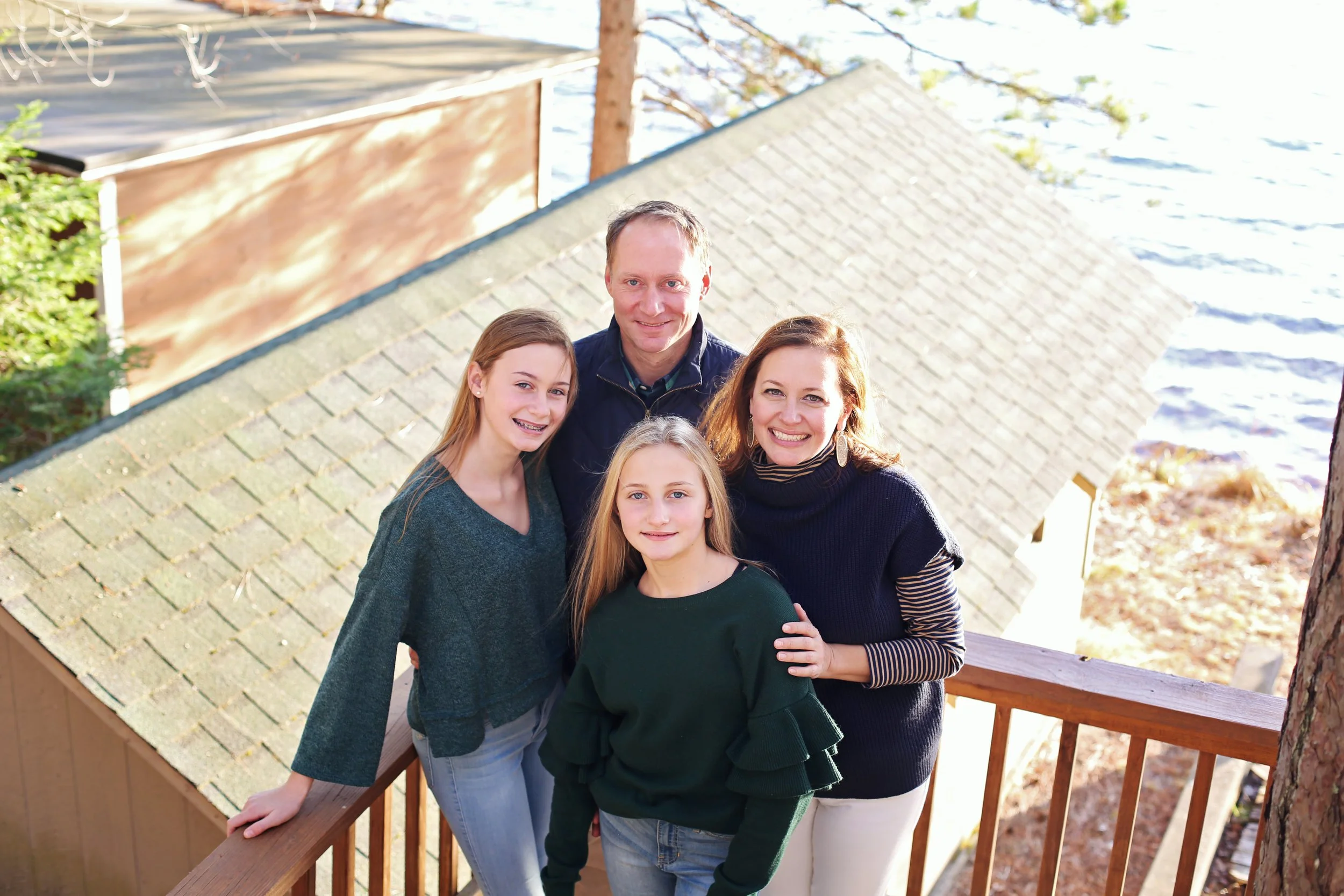 A group of four people, including two adults and two children, standing on a wooden deck outdoors near a lake, smiling and looking up at the camera.