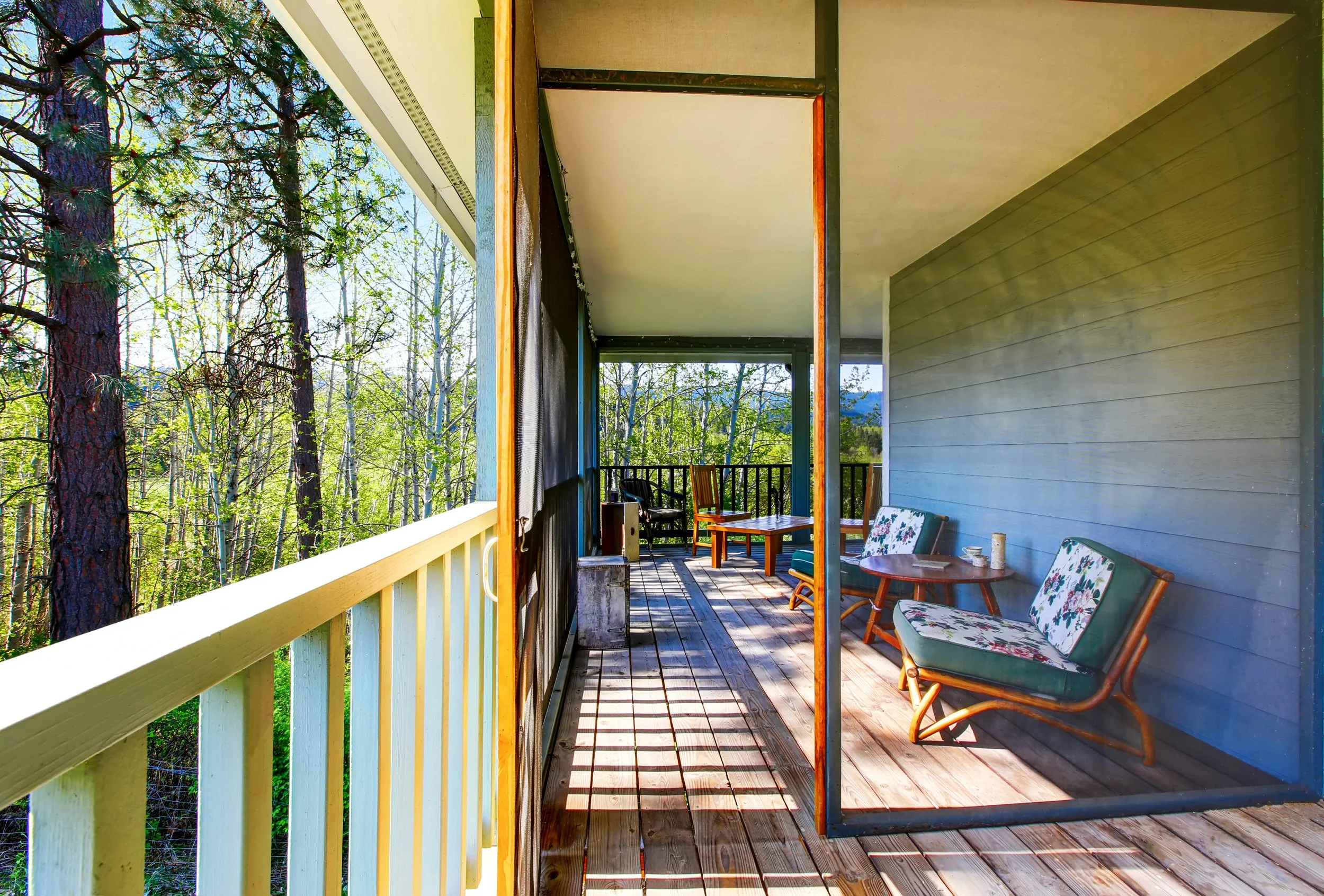 A covered porch with wooden flooring and chairs, with a view of trees and sky in the background.