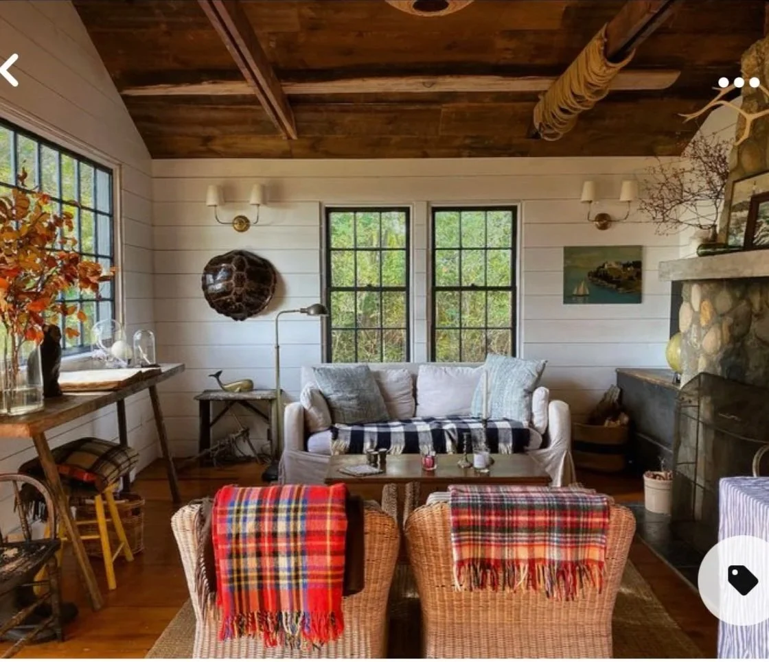 Cozy living room with white wooden wall panels, large black-frame windows, a stone fireplace, a cream sofa with plaid and plain pillows, two wicker chairs with red plaid blankets, a wooden coffee table, and various decorative items including dried flowers and artwork.