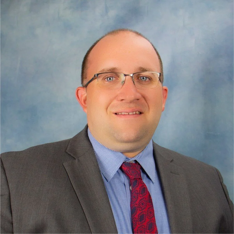 A man in a suit and tie with glasses posing for a professional headshot against a blue background.
