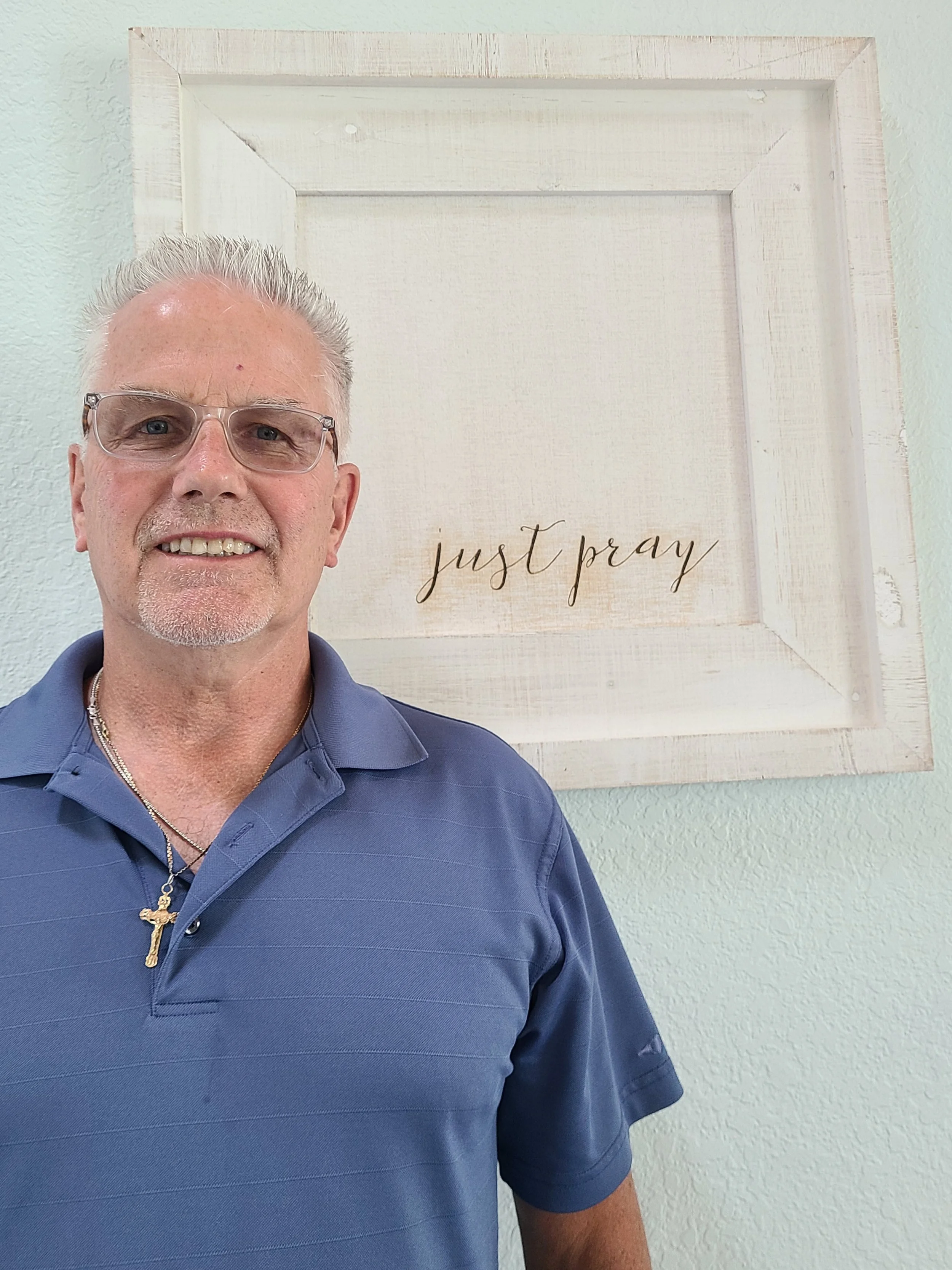A man with gray hair, glasses, and a goatee wearing a blue polo shirt with a gold cross necklace, standing in front of a white framed art piece with the words "just pray" written in script.