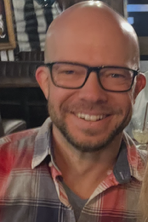 A smiling man with glasses and a beard, wearing a plaid shirt in a cozy indoor setting.