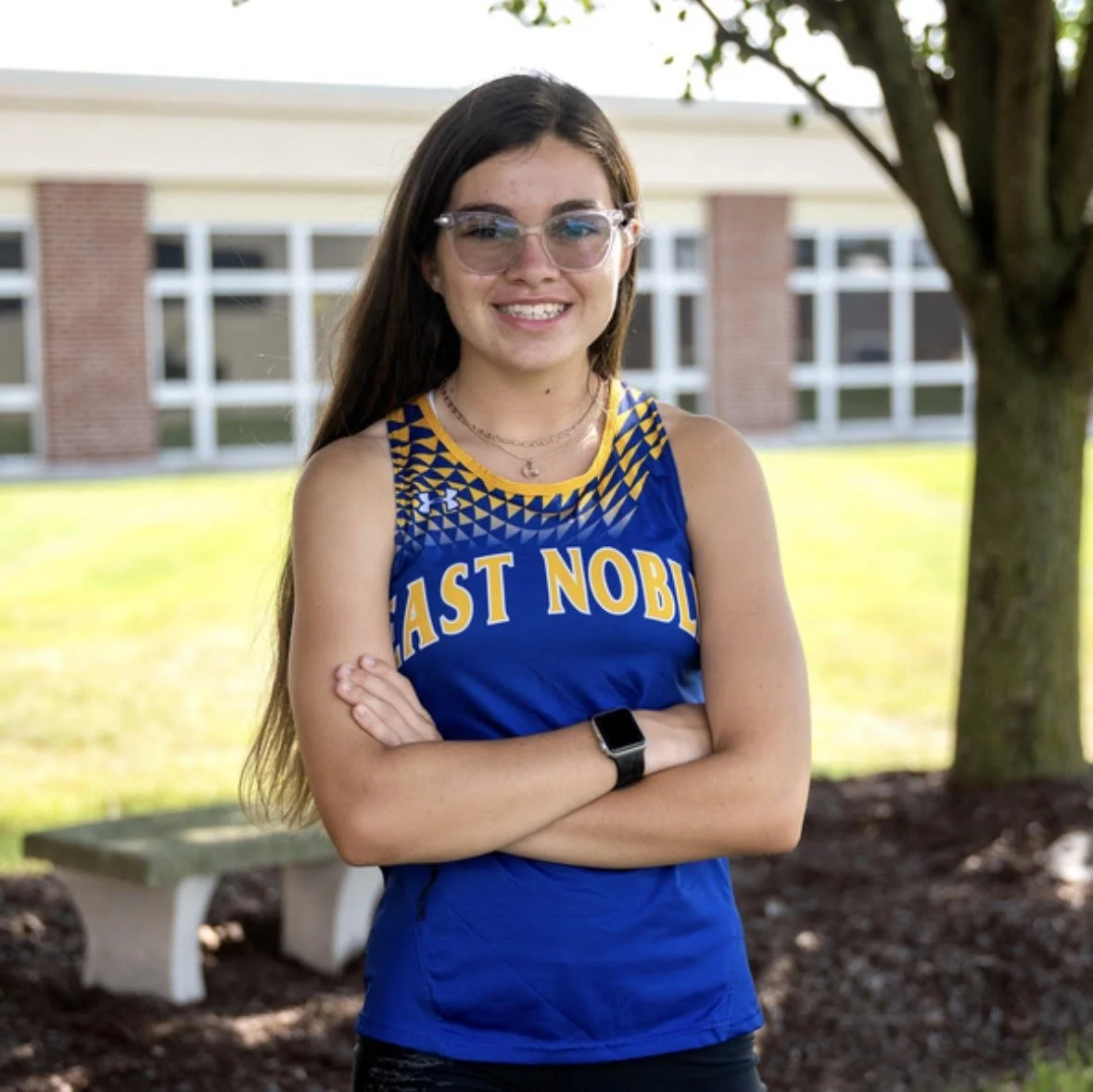 A young woman with long brown hair wearing glasses, a blue and yellow East Noble track uniform, and a black smartwatch, standing outdoors with arms crossed and smiling.