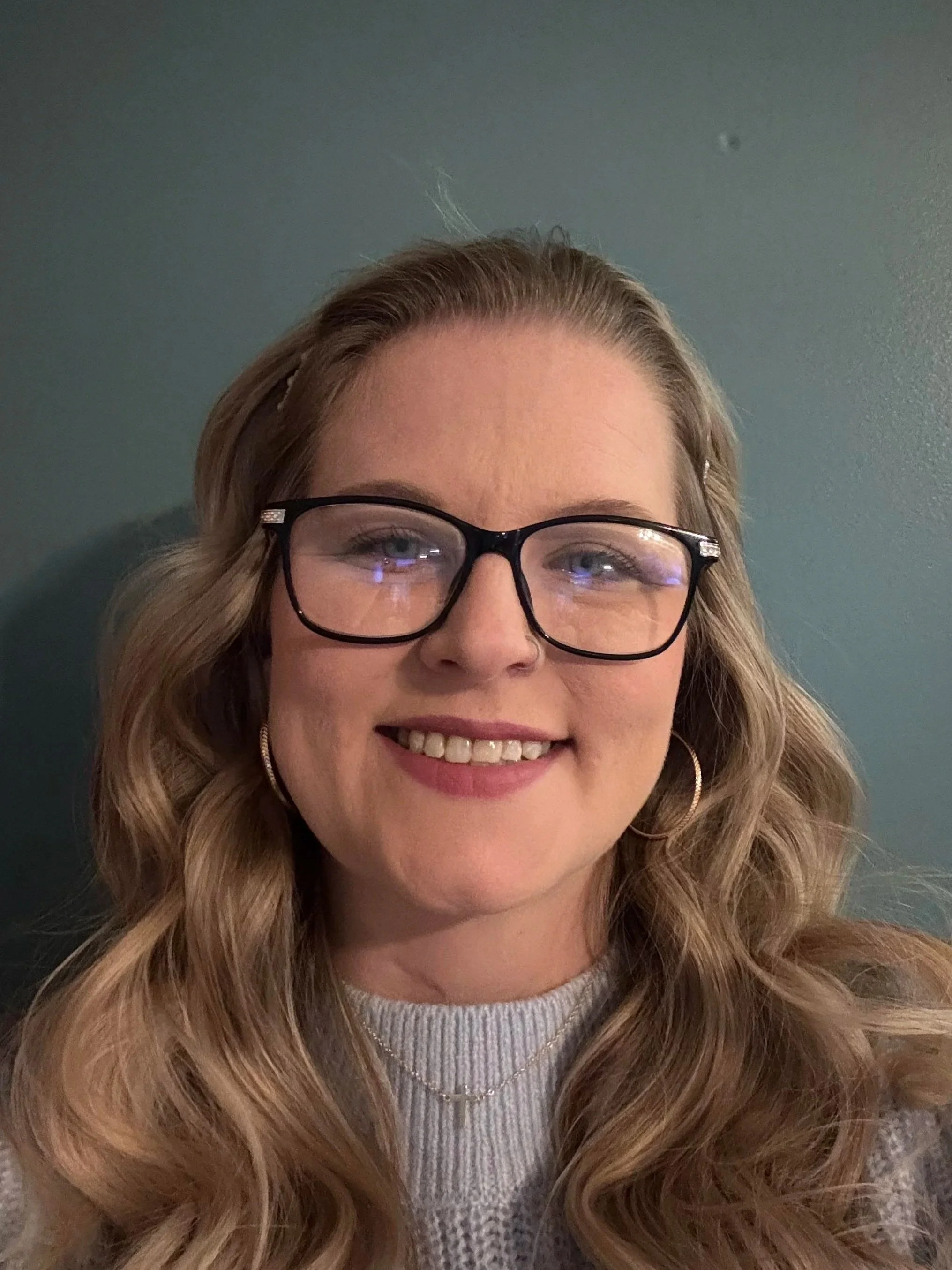 A woman with long wavy blonde hair wearing glasses, a light gray knit sweater, and a white top, smiling in front of a blue wall.