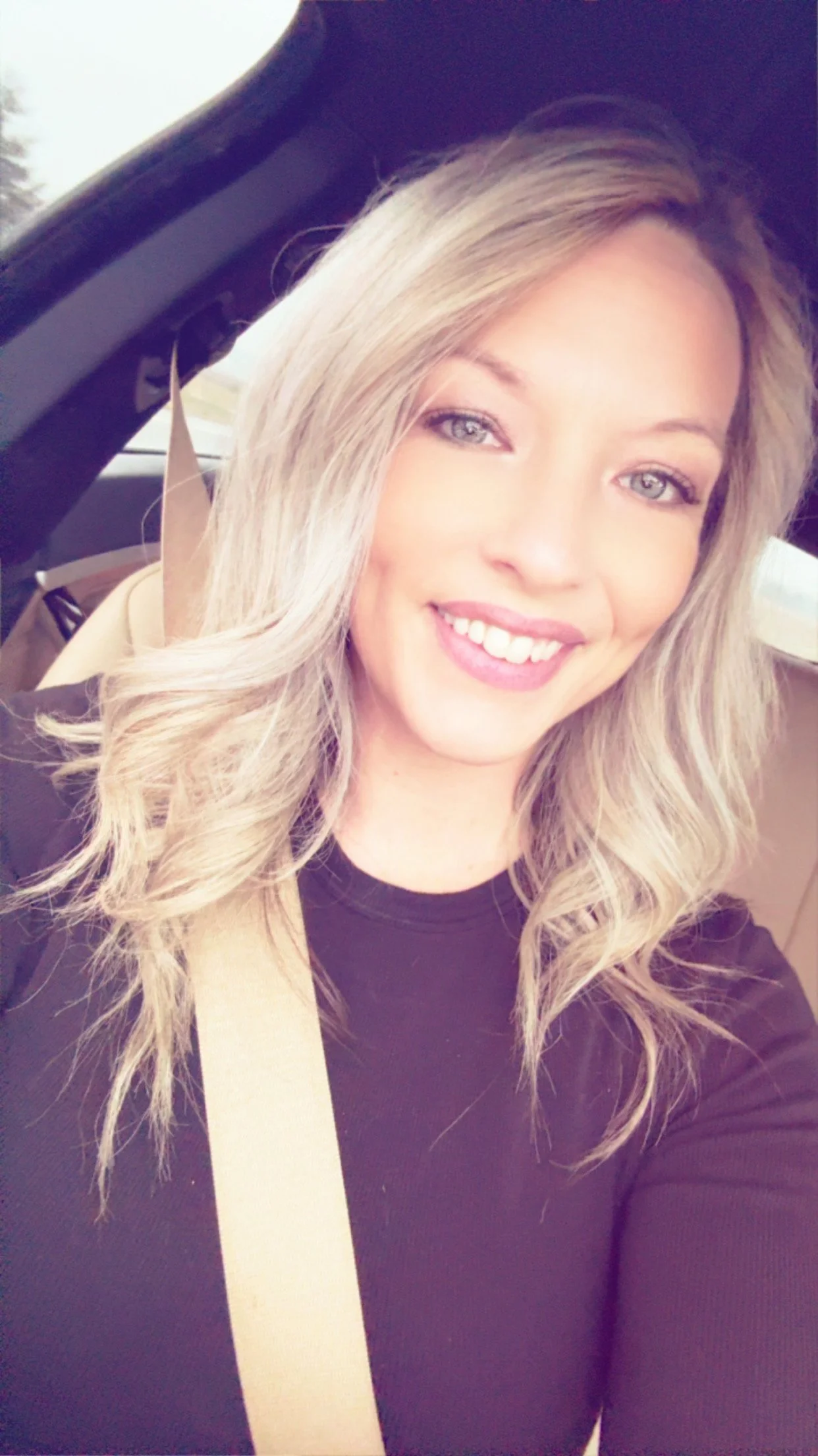 A woman with long blonde hair and blue eyes smiling while sitting in a car, wearing a black top and a seatbelt.