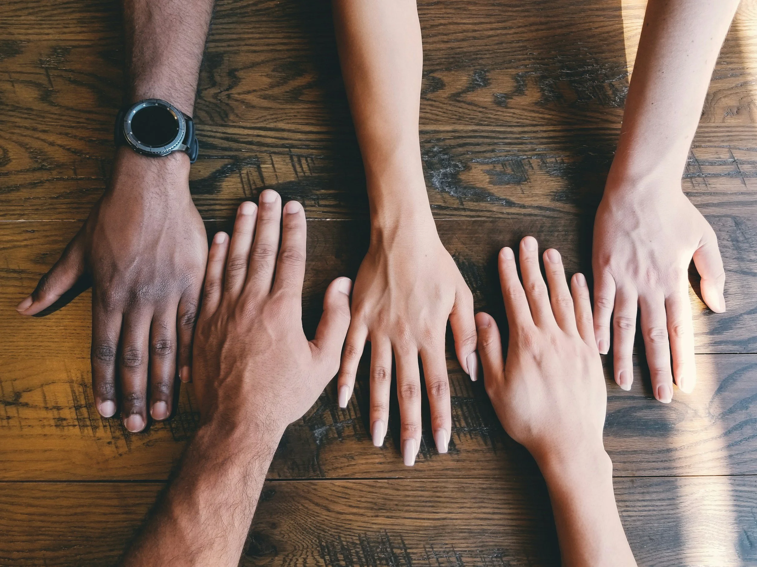 Multiple hands, diverse in skin tones, placed on a wooden table, with a person's wristwatch visible on one wrist.