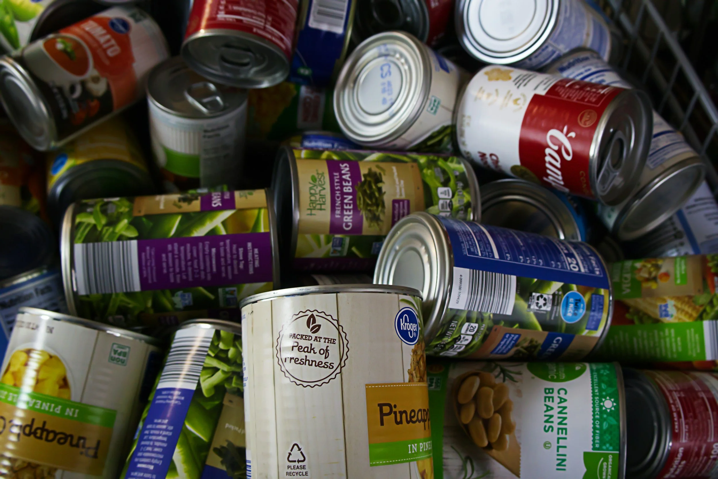 A pile of various canned foods and boxed products, including green beans, pineapple chunks, and other vegetables, in a grocery cart.