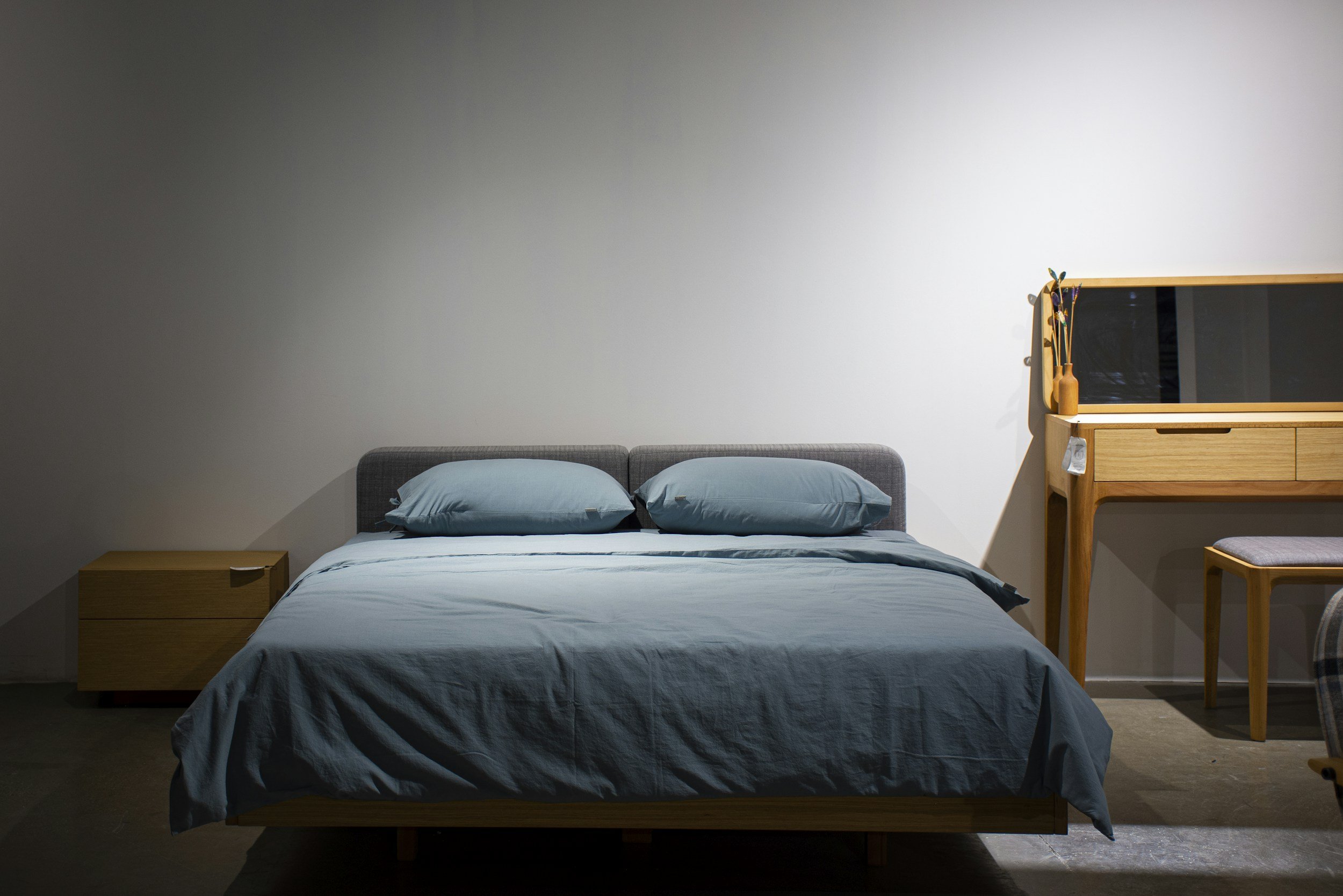 A neatly made bed with a dark gray headboard, light blue bed linen, and matching pillows. To the left, a small wooden nightstand. To the right, a wooden dresser with a mirror, a dried plant, and decorative items on top. The room has a plain white wall background.