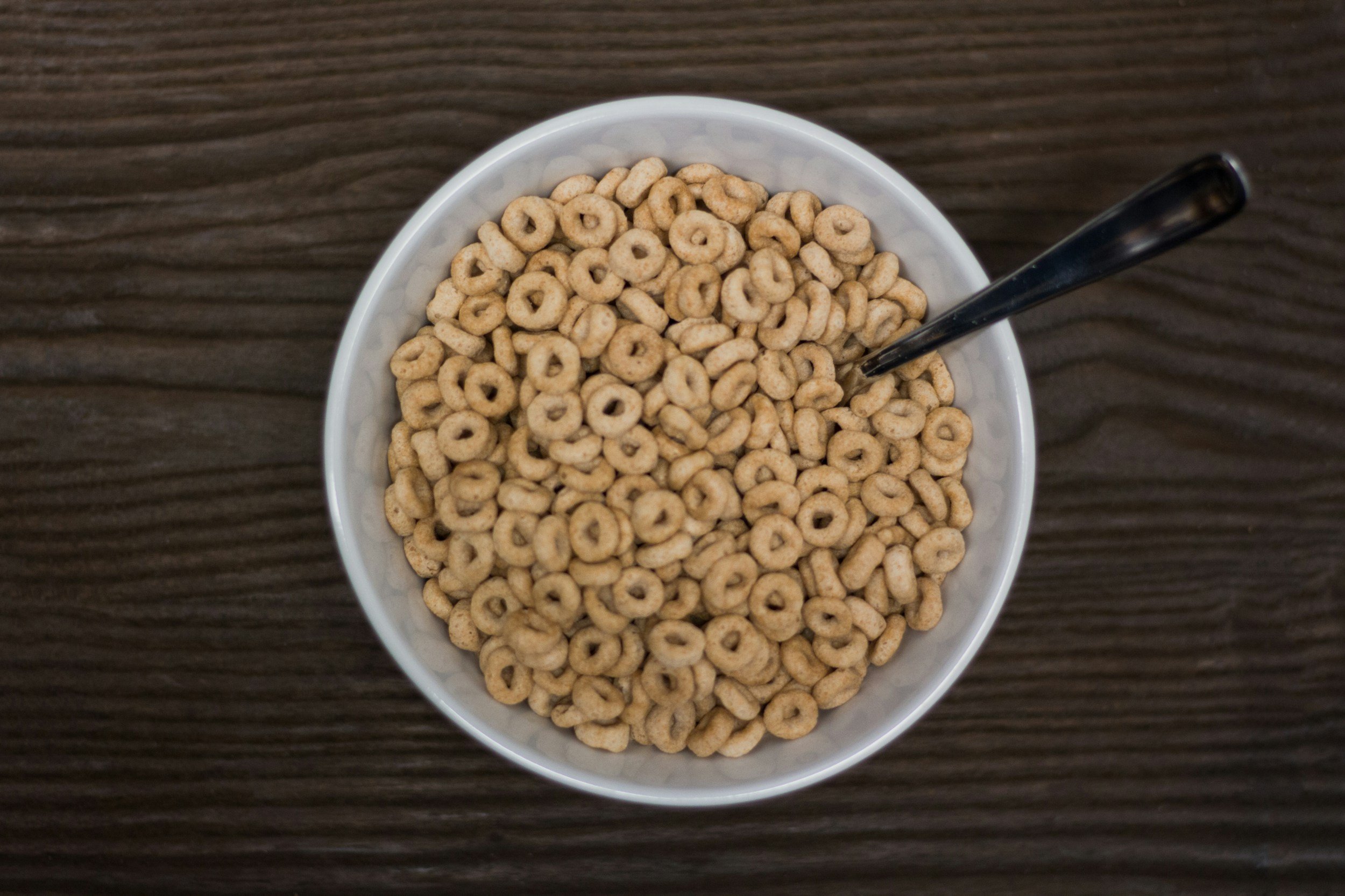 A white bowl filled with small, round cereal pieces on a dark wooden surface, with a black spoon resting inside the bowl.