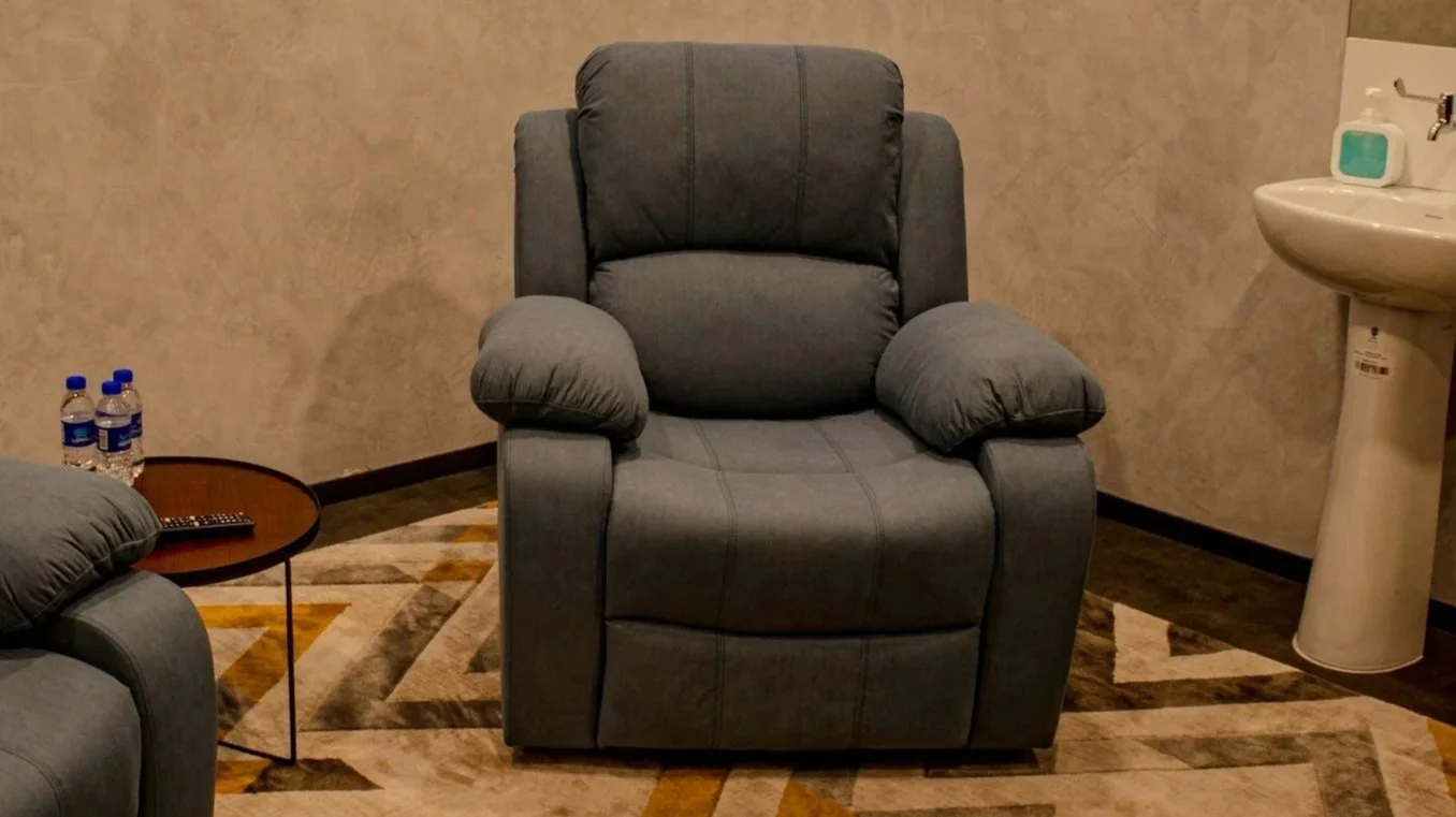 Gray recliner chair in a room with beige walls, a small round side table holding two bottled waters and a remote, a patterned rug on the floor, and a white sink with a soap dispenser in the background.