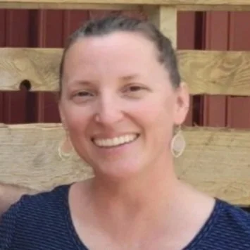 A woman with light skin and dark hair pulled back, smiling, wearing a navy blue top and earrings, standing in front of a wooden fence.
