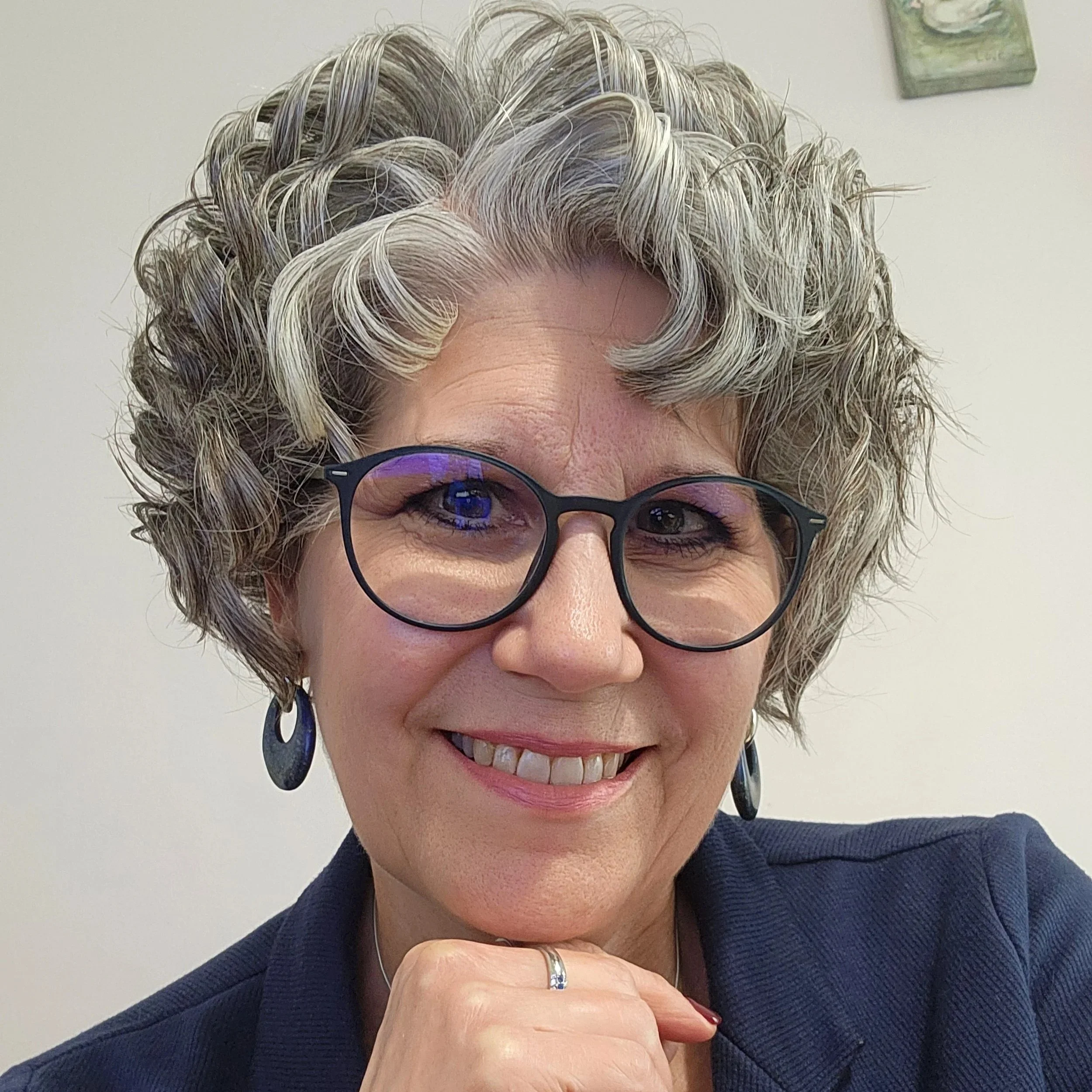 A woman with short, curly, gray hair, wearing glasses with dark frames, a dark blazer, and earrings, smiling.