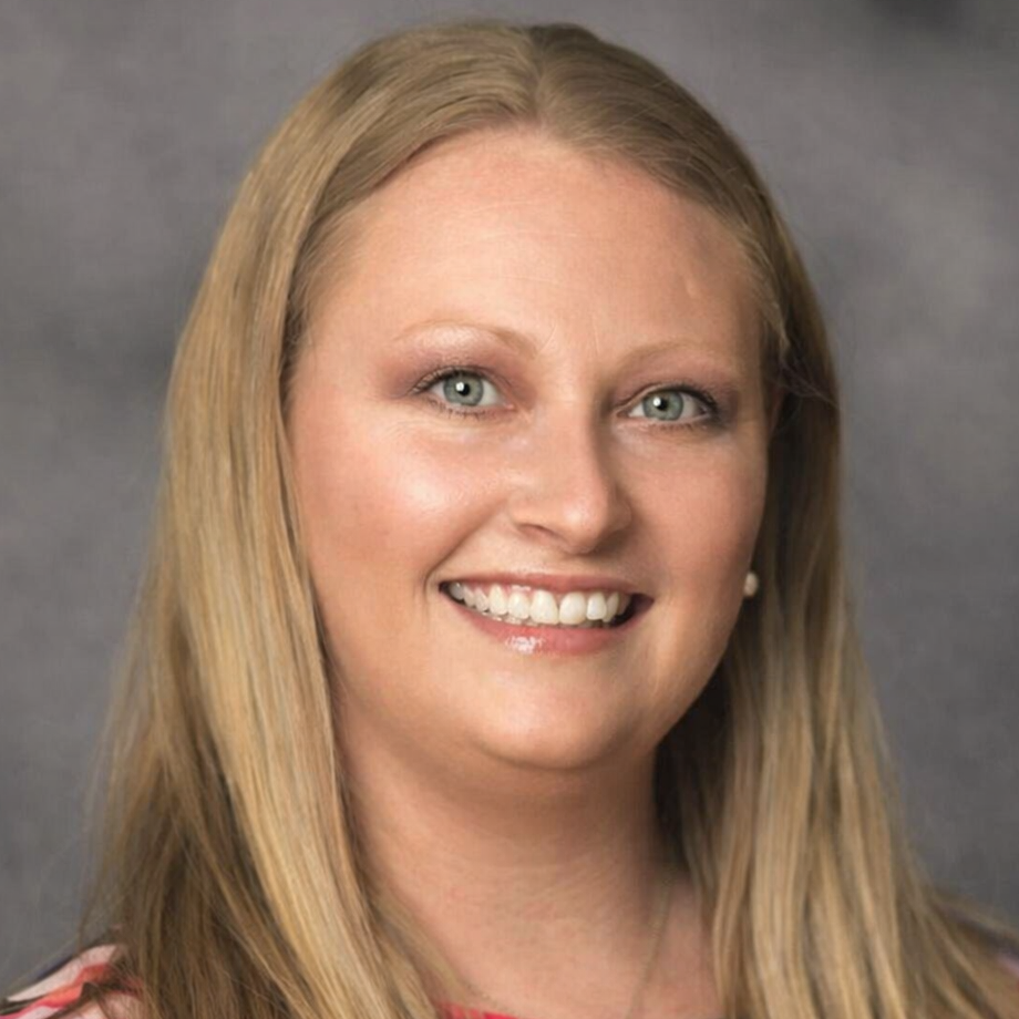 A portrait of a smiling woman with long blonde hair, blue eyes, wearing a pink top and pearl earrings, against a gray background.
