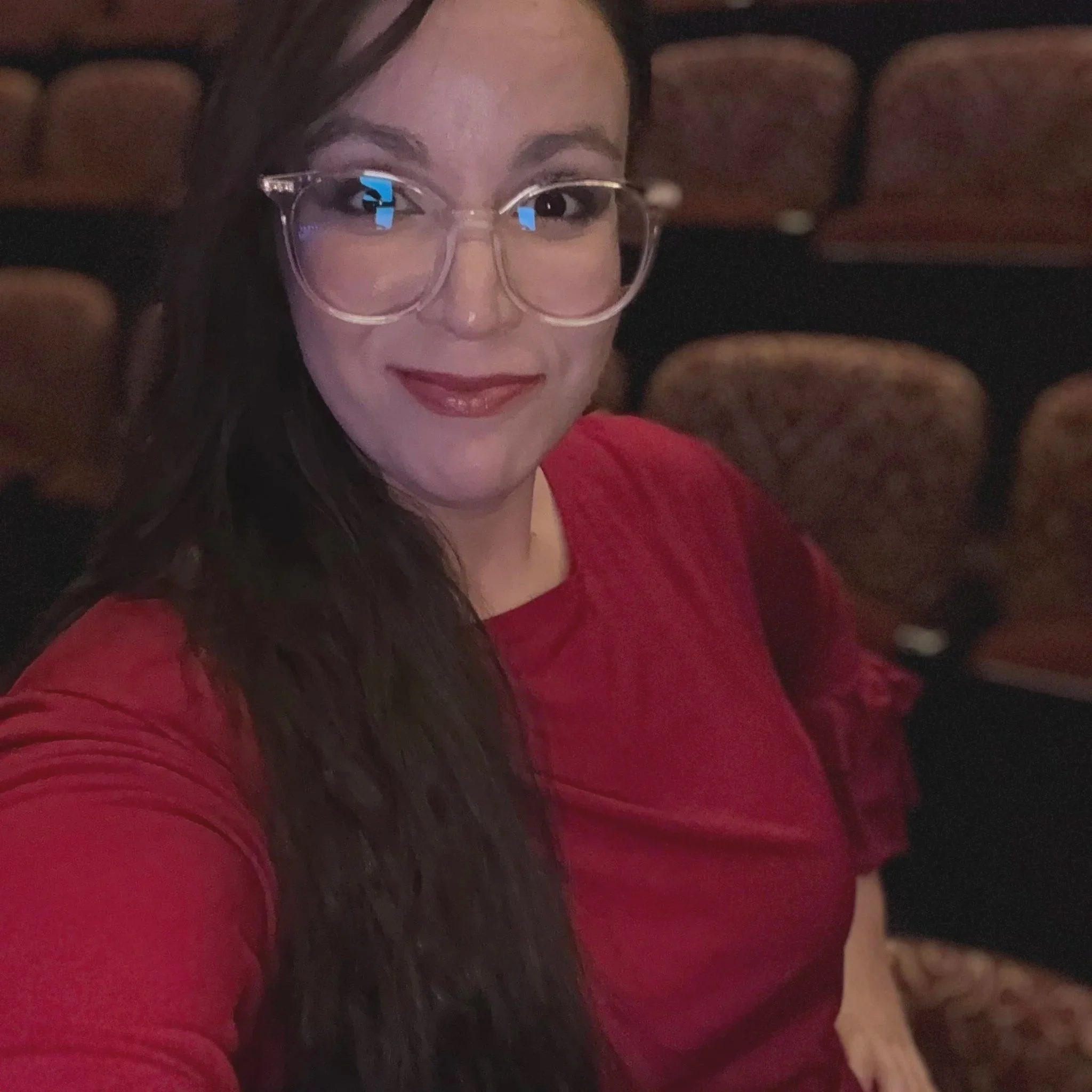 A woman with long dark hair, wearing glasses and red lipstick, smiling while holding a small dog with brown and white fur, on a brown leather couch.