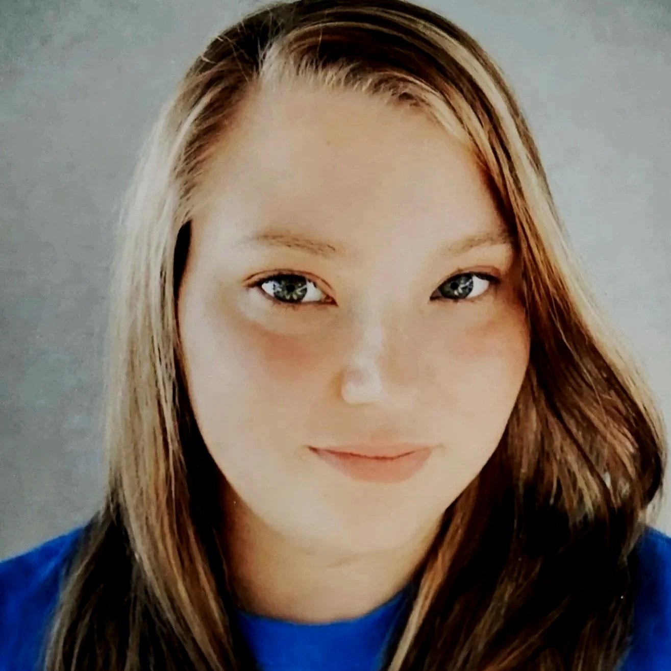 Close-up of a woman with long, light brown hair and blue eyes, wearing a blue shirt, looking at the camera against a light gray background.