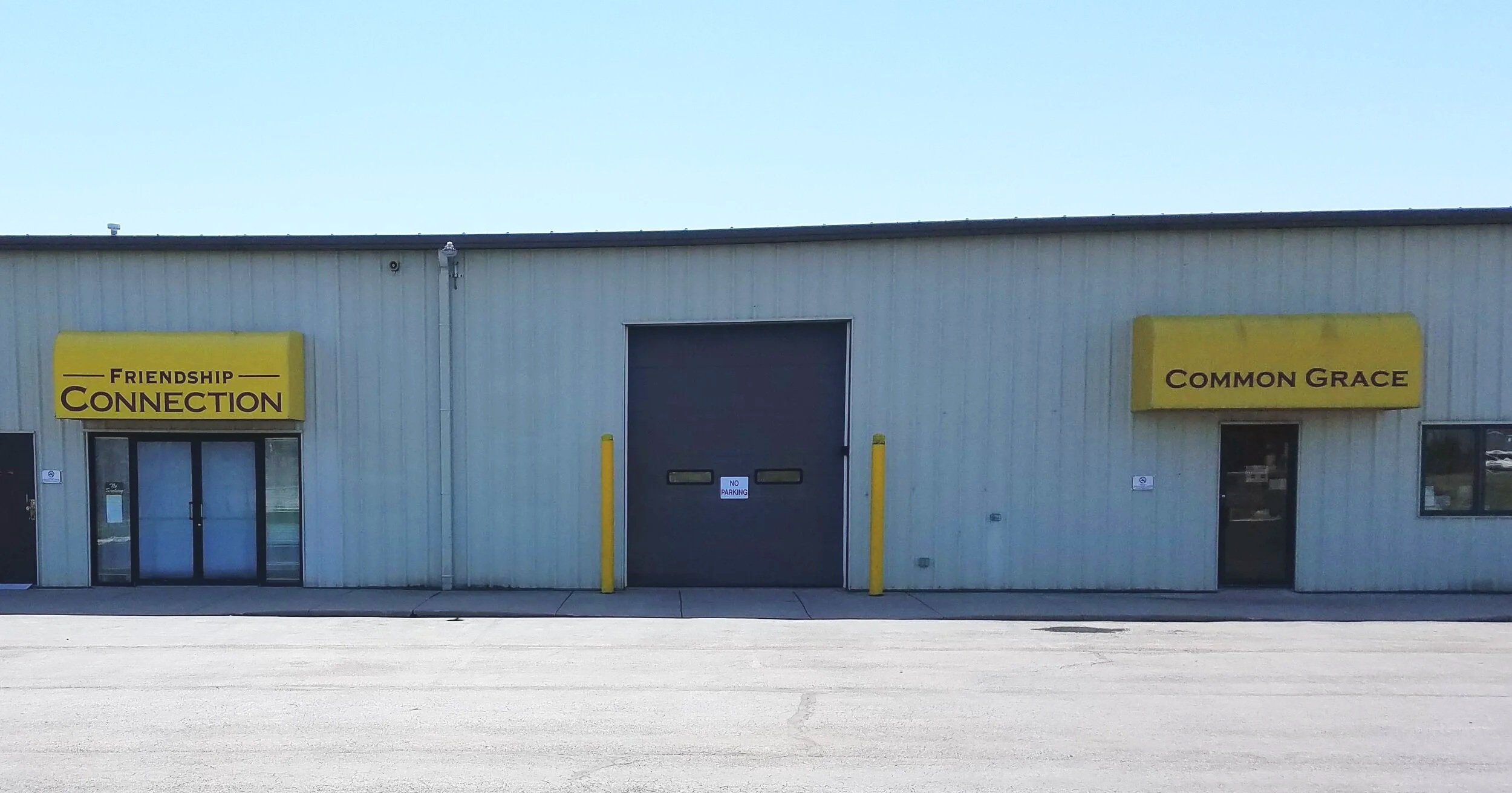 A beige industrial building with two yellow signs that read 'Friendship Connection' and 'Common Grace'. There are glass doors and a large black door with a sign that reads 'No Parking'.